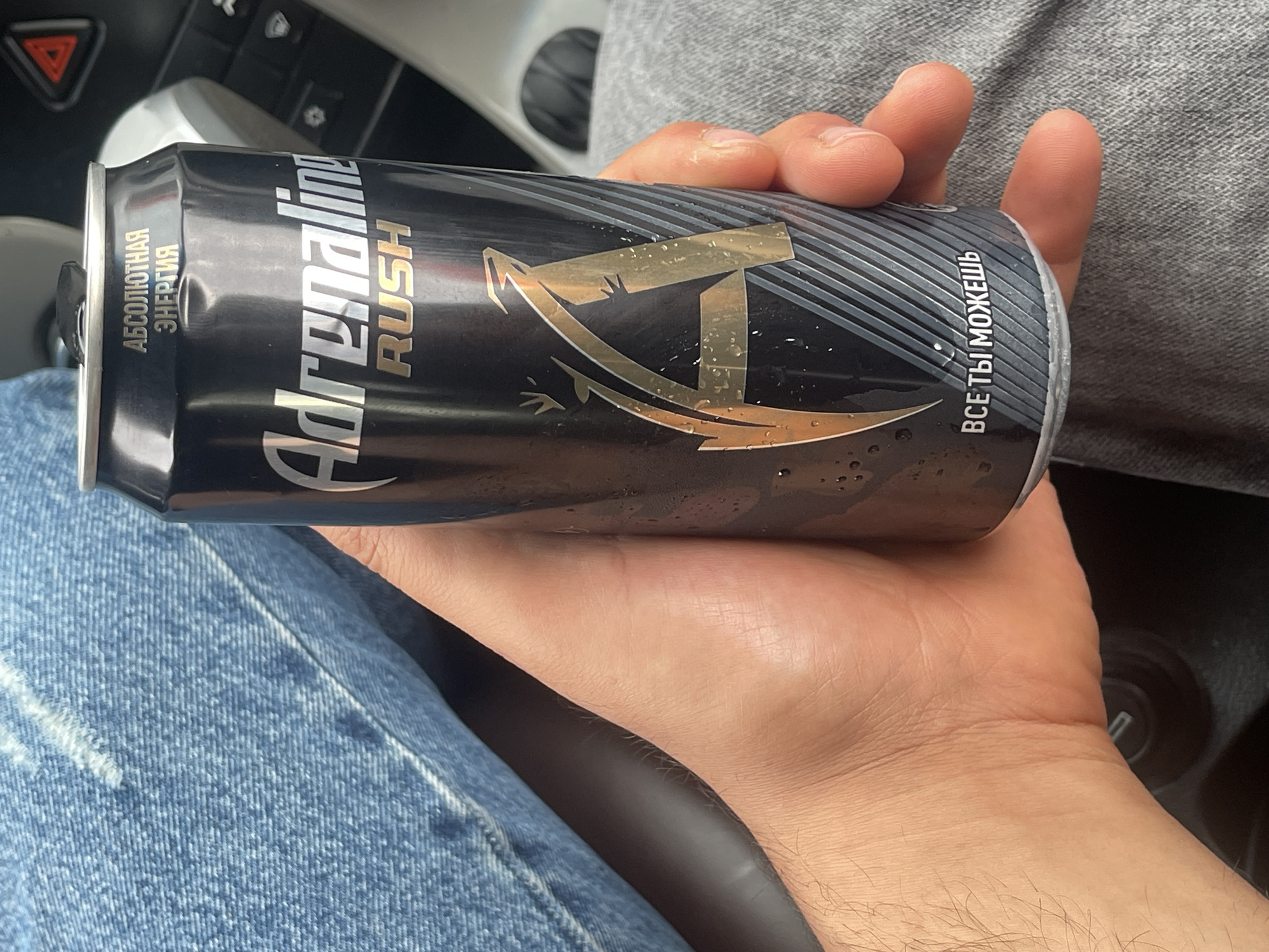 In the city of Istra, Russia, a person's hand holds a black can of "Adrenaline Rush" energy drink. The can has a large golden "A" logo and text in Russian that reads "АБСОЛЮТНАЯ ЭНЕРГИЯ" (Absolute Energy) and "ВСЕ ТЫ МОЖЕШЬ" (You Can Do Anything). The background shows the person's legs, clad in blue jeans and grey sweatpants, and a car's interior, including a red triangular warning light. The can appears to be slightly condensed.