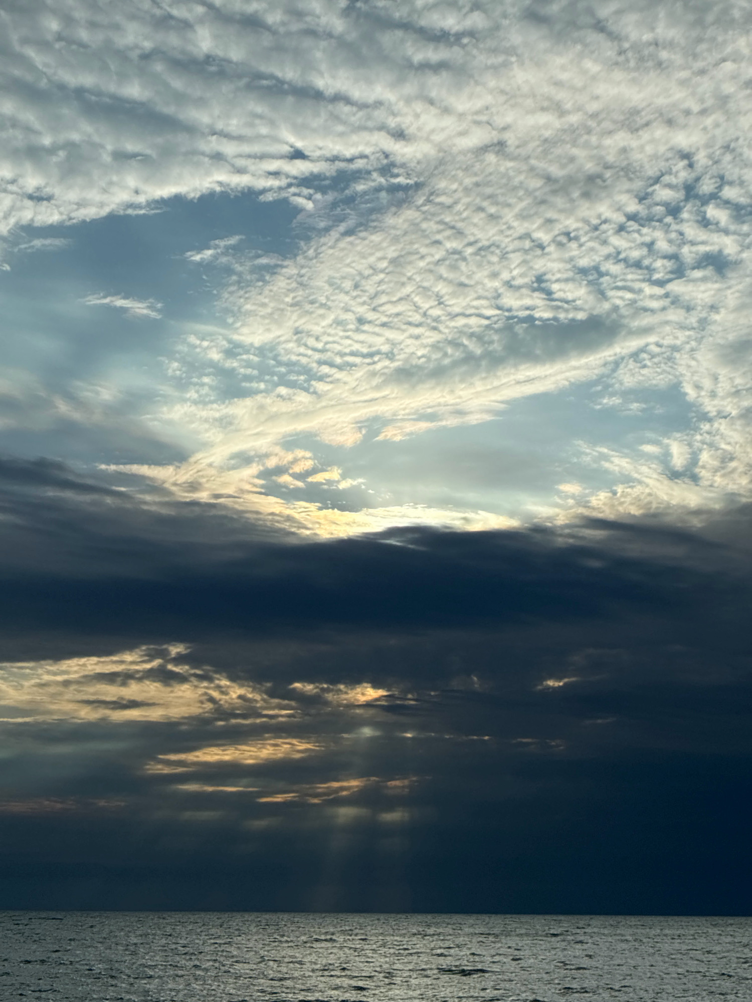The image depicts a dramatic sky over the Black Sea, viewed from the coast of Sochi, Russia. The sky is filled with a mix of dark, heavy clouds and lighter, wispy formations, with beams of sunlight breaking through in several areas. The sea is visible in the foreground, appearing choppy and reflecting the muted light. The overall impression is one of a dynamic and somewhat stormy atmosphere. There are no people or man-made structures visible in the frame.