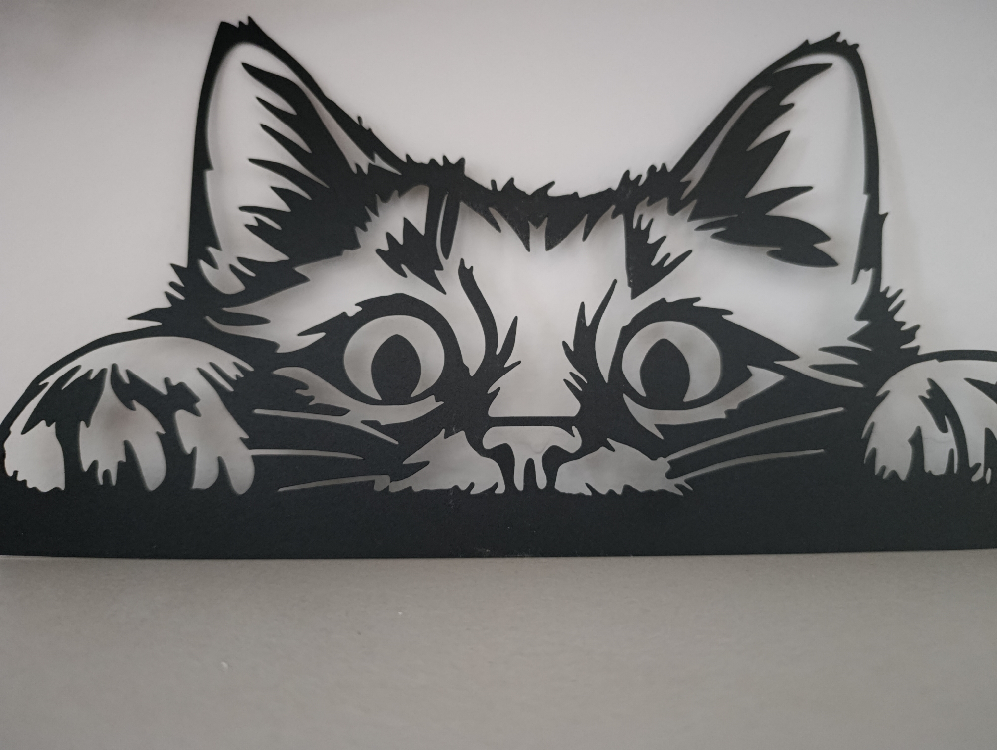 A close-up shot shows a black, cutout metal artwork of a cat's face and paws. The cat's eyes are wide and expressive. The ears are tall and pointed. The paws are slightly visible at the bottom left and right. The artwork is placed against a white and gray background. The location is Unknown, Unknown.