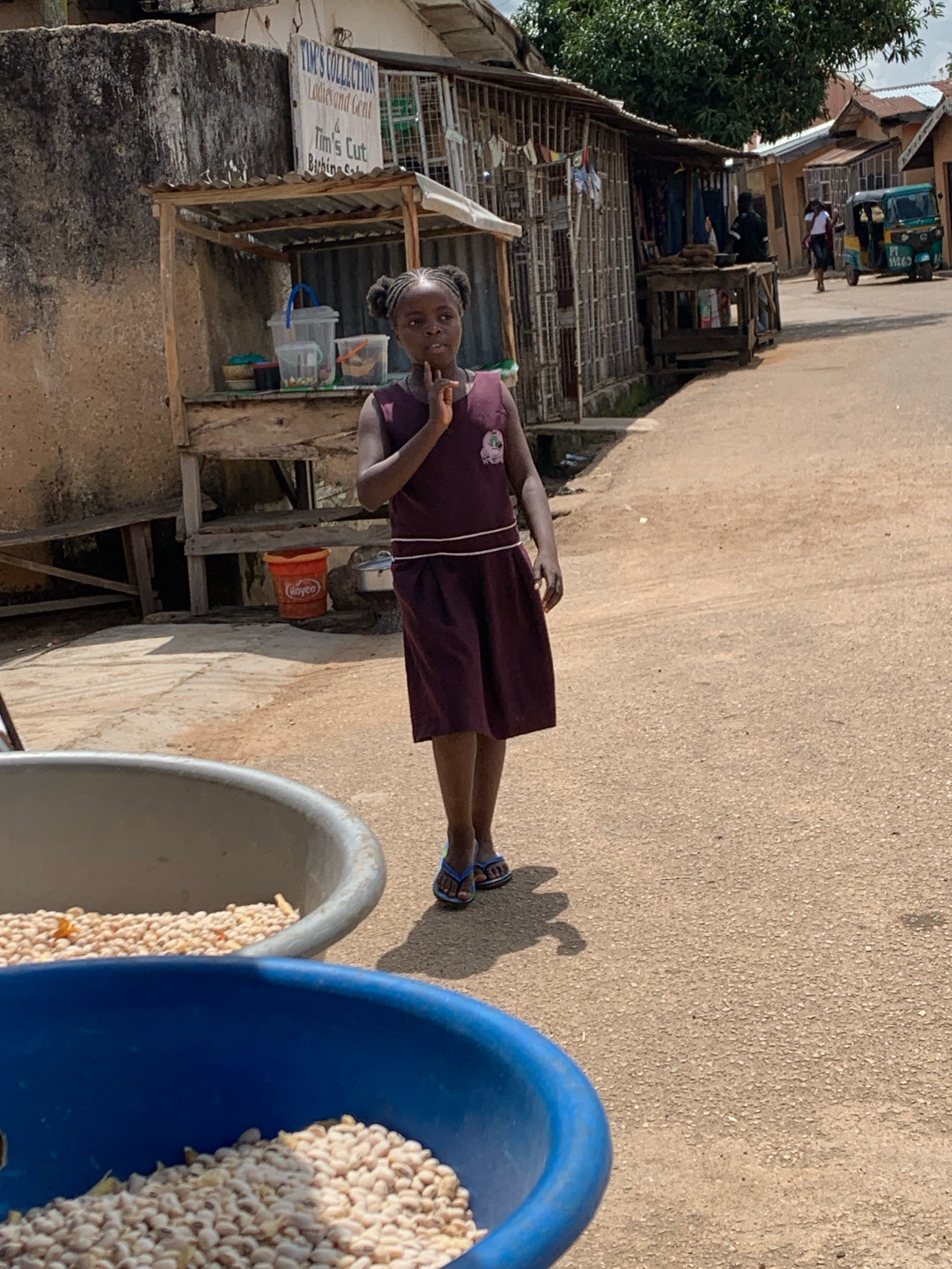 A young Black girl with her hair in two pigtails stands on a dusty road in front of a market stall in Jos, Nigeria. She is wearing a maroon dress and blue flip-flops and has her hand raised to her chin in a thoughtful pose. The market stall behind her is made of wood and corrugated metal and is filled with various goods. In the foreground, two large bowls, one gray and one blue, are filled with what appears to be dried beans or peas. In the background, other market stalls and buildings line the road, and a three-wheeled vehicle is parked nearby. The overall scene depicts a bustling outdoor market in an African city.