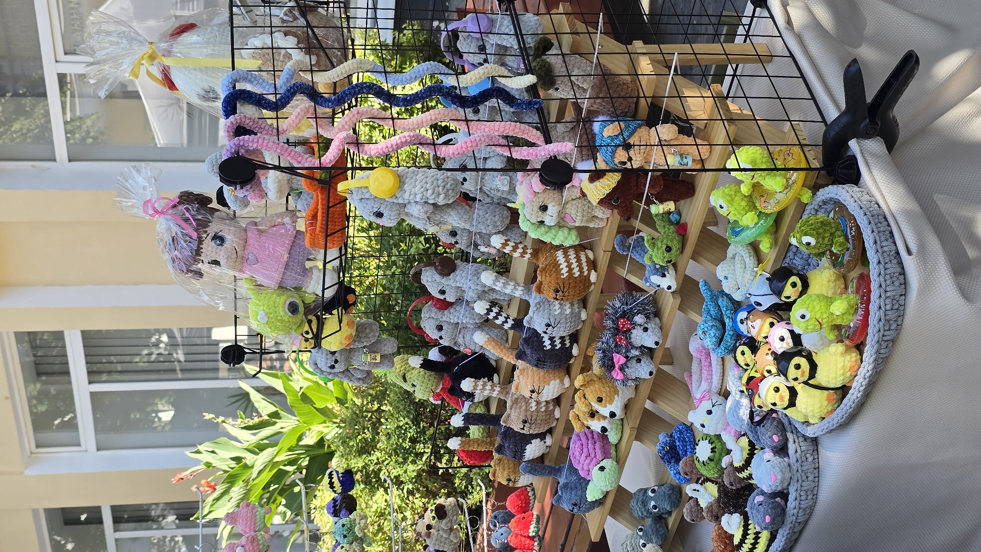 The image depicts an outdoor stall displaying a large assortment of crocheted stuffed animals, primarily in various animal forms and some doll-like figures. The stall is set up on a table with a light-colored tablecloth. Behind the table, a wooden tiered display rack holds many of the crocheted items. The background shows green foliage and the facade of a building with windows, suggesting an outdoor market or fair.
Specifically, the stall is located in the city of Zhytomyr, Ukraine. The assortment includes numerous small crocheted animals like cats, dogs, hedgehogs, frogs, and birds, some of which are arranged in baskets or trays. Larger crocheted items, such as dolls and what appear to be rabbits or other long-eared creatures, are displayed on the tiered rack and on the top of the stall. Many of these items are vibrant and colorful. The lighting suggests it is daytime, likely under natural sunlight. There are no people clearly visible in the frame interacting with the items. The overall scene is one of a vendor's display of handmade crafts.