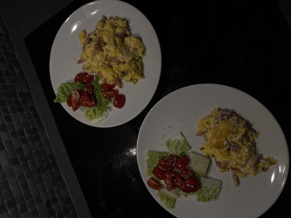 This image captures a domestic kitchen scene in Warsaw, Poland. Two identical meals are presented on white, round plates, resting on a black, reflective induction cooktop, which bears a "BOSCH" logo in the lower right quadrant. The background reveals a dark, small-square tiled backsplash extending upwards from a dark counter edge. Each plate features a serving of scrambled eggs intermixed with diced ham or bacon and green chives. Adjacent to the eggs, a side salad consists of chopped Napa cabbage leaves and halved red cherry tomatoes, with several tomatoes garnished with white sesame seeds. No individuals are present, and no active interactions are depicted.