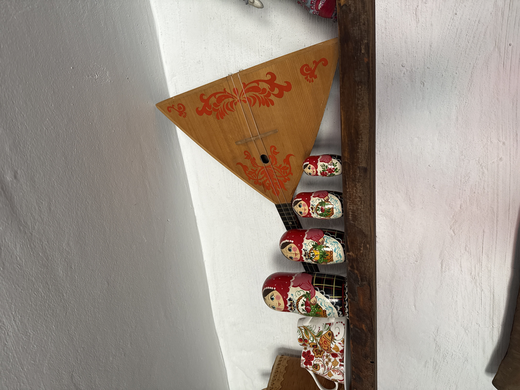On a wooden shelf, several Russian nesting dolls and a balalaika are displayed. The dolls are of varying sizes, painted with traditional floral patterns and faces. The balalaika, a triangular stringed instrument, leans against the wall behind the dolls and is decorated with red floral motifs. Two patterned mugs are visible to the left of the dolls. The shelf is mounted on a white, textured wall, and a white ceiling is visible above. This scene appears to be from a home or shop in Noviy, Russia, likely showcasing traditional Russian crafts.