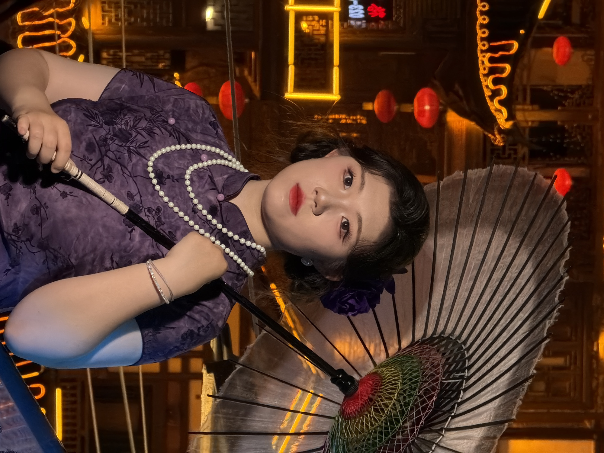A young East Asian woman with dark hair styled in a bob, wearing a purple floral print qipao, a multi-strand pearl necklace, and a floral hair accessory, holds a traditional Japanese umbrella. Her makeup is notable with bold red lipstick and defined eyeliner. The background suggests an outdoor setting at night in what appears to be a historic district with traditional Chinese architecture, illuminated by red lanterns and yellow neon lights, possibly in Yuzhong District, China. The lighting casts warm tones on the scene, highlighting the textures of the woman's attire and the intricate details of the umbrella. The woman's gaze is directed towards the viewer, with a neutral expression.