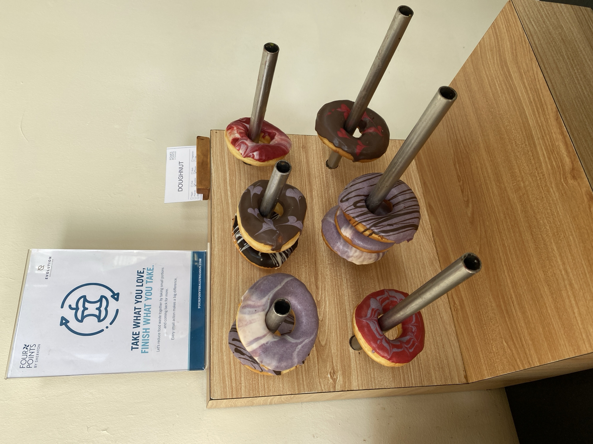 The image depicts a breakfast buffet display at the Four Points by Sheraton Bali, Ungasan, located in Kuta Selatan, Indonesia. The main subject is a wooden stand holding several doughnuts on metal rods. The doughnuts are ring-shaped and decorated with various glazes, including purple, chocolate, and red swirled icing. A sign above the doughnuts on the left promotes a "Take What You Love, Finish What You Take" initiative aimed at reducing food waste. To the right of this sign, a smaller card labeled "DOUGHNUT" with checkboxes for dietary information is partially visible. The background is a plain wall and a wooden surface. The scene appears to be indoors, likely in a hotel restaurant or breakfast area.