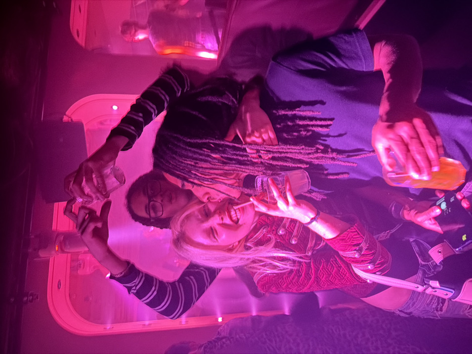 Three people are in a dimly lit setting, likely a bar or club. A blonde woman in a red jacket smiles, holding a drink and a lollipop. Behind her, a person with dreadlocks is partially visible. Another individual wearing glasses and a striped shirt holds a glass. The scene is bathed in a pink and purple light. A couch and a bar counter with bottles on display are visible in the background.
