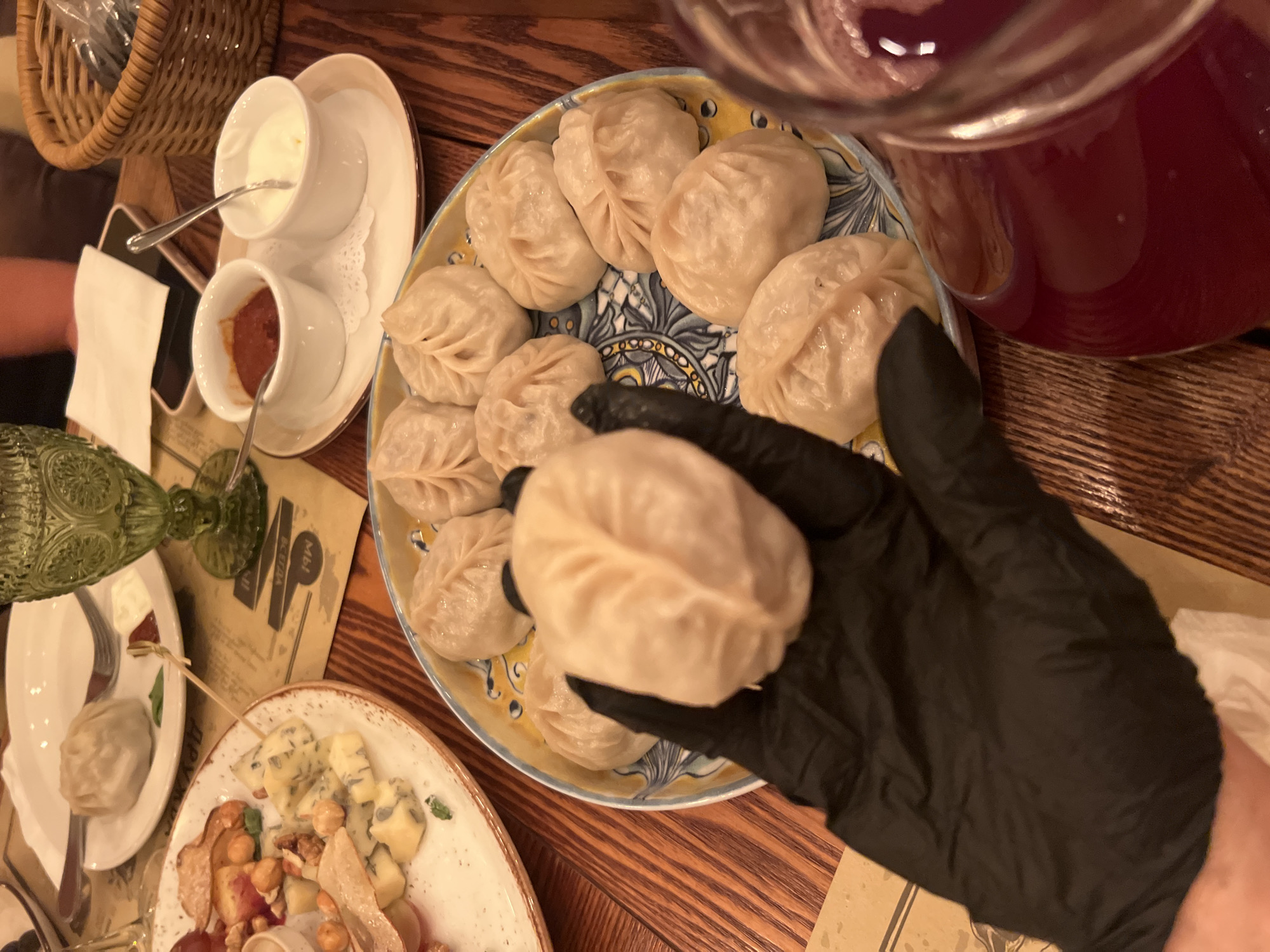 In this close-up, overhead shot, a hand clad in a black disposable glove is holding a single, pale dumpling above a patterned blue and yellow ceramic platter filled with other identical dumplings. The dumplings appear to be steamed, with a smooth, pleated surface. The platter is set on a wooden table. In the background, out of focus, are other plates of food, a green glass goblet, two small white bowls containing red sauce and a white cream or yogurt, and a woven basket. The setting appears to be a restaurant.