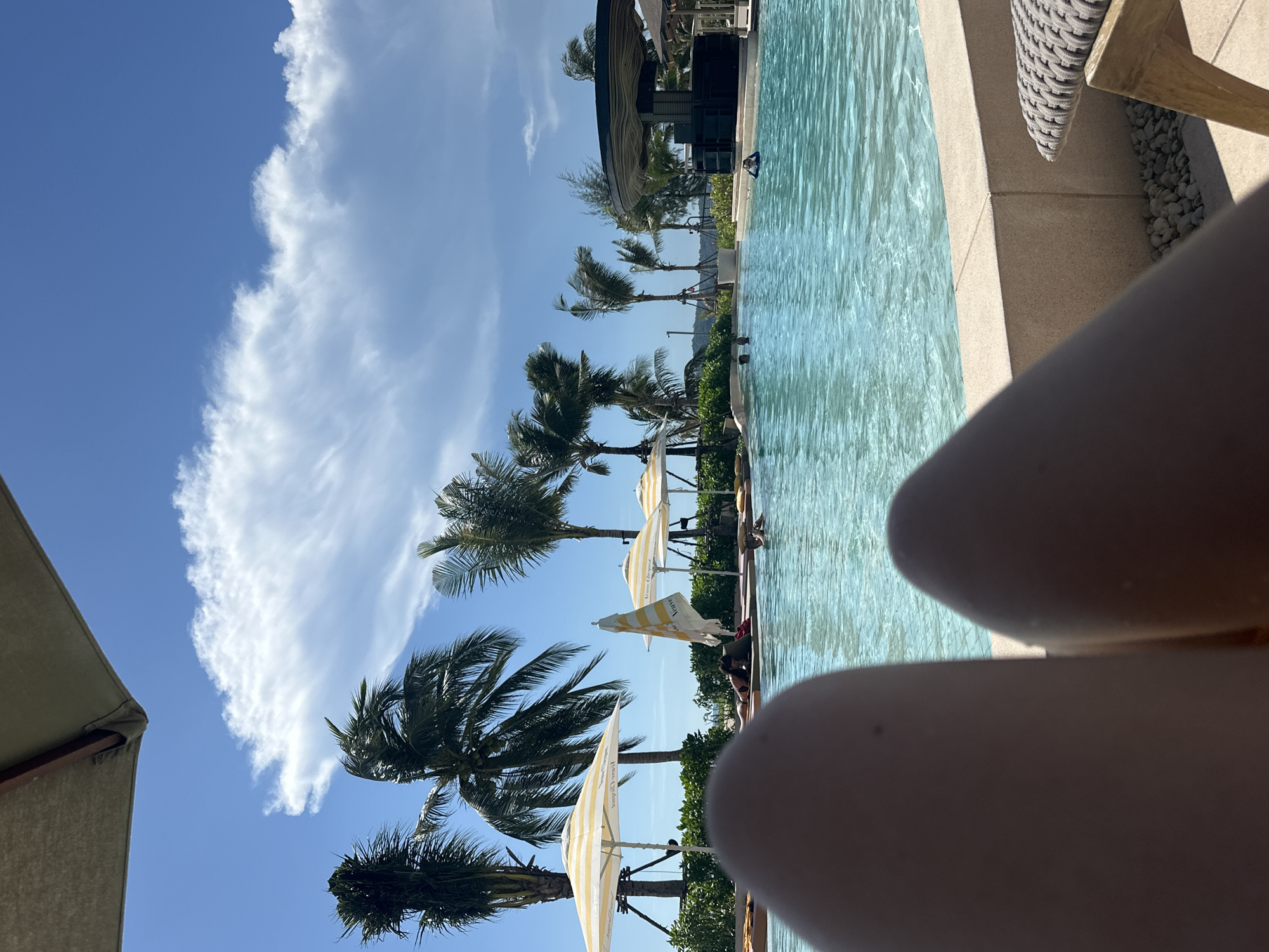 In a resort setting, the camera is positioned at a low angle, looking over the legs of an individual relaxing poolside. The foreground is dominated by the blurred shapes of these legs, suggesting the viewer is lying down on a lounge chair. Beyond them, a bright blue swimming pool stretches out, its surface rippling under the sunlight. Several people are visible in the pool and around its edge. Palm trees line the background, some swaying gently in the wind, with beach umbrellas providing shade. A circular bar structure is visible to the right of the pool. The sky is a vibrant blue, dotted with fluffy white clouds. The overall scene evokes a vacation atmosphere.