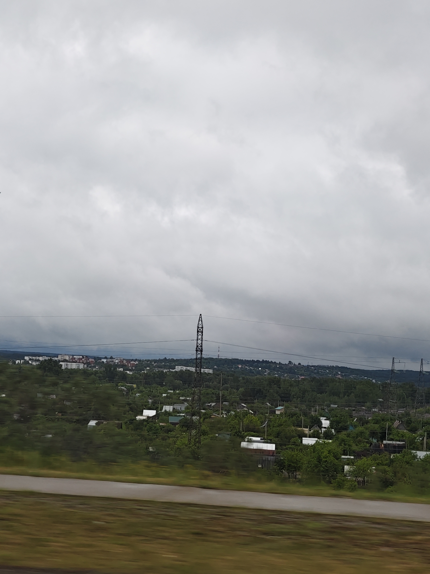 The image depicts a landscape near Chaikovsky, Russia, under a cloudy sky. A distant town with apartment buildings is visible on a hill. In the mid-ground, there is a mix of trees and small structures that appear to be houses or dachas (country houses) interspersed with trees. Power line pylons are visible. The foreground shows a blurred field and road. The photo seems to have been taken from a moving vehicle, possibly a train or car.