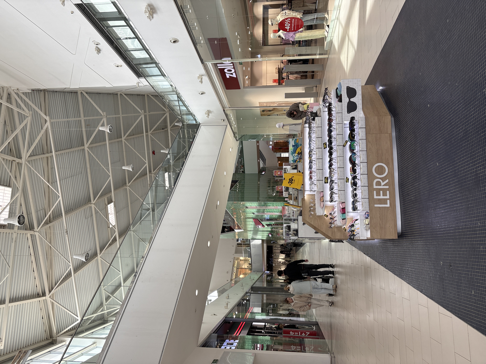 The image captures the interior of a shopping mall, likely located in Ulyanovsk, Russia, based on the Cyrillic text visible in the store names and promotional signs. The environment is bright and spacious, characterized by high ceilings with exposed steel beams and skylights. A multi-level design incorporates glass barriers and escalators to connect different floors.
People are present, including shoppers browsing in front of a "LERO" sunglasses display stand with "30%" off signs. Nearby are retail stores, one with a "zoll" sign and another with a promotion indicating "-40% on the second item." The flooring is a mix of light and dark tiles. The camera angle provides a slightly elevated perspective, looking down the mall's central walkway.