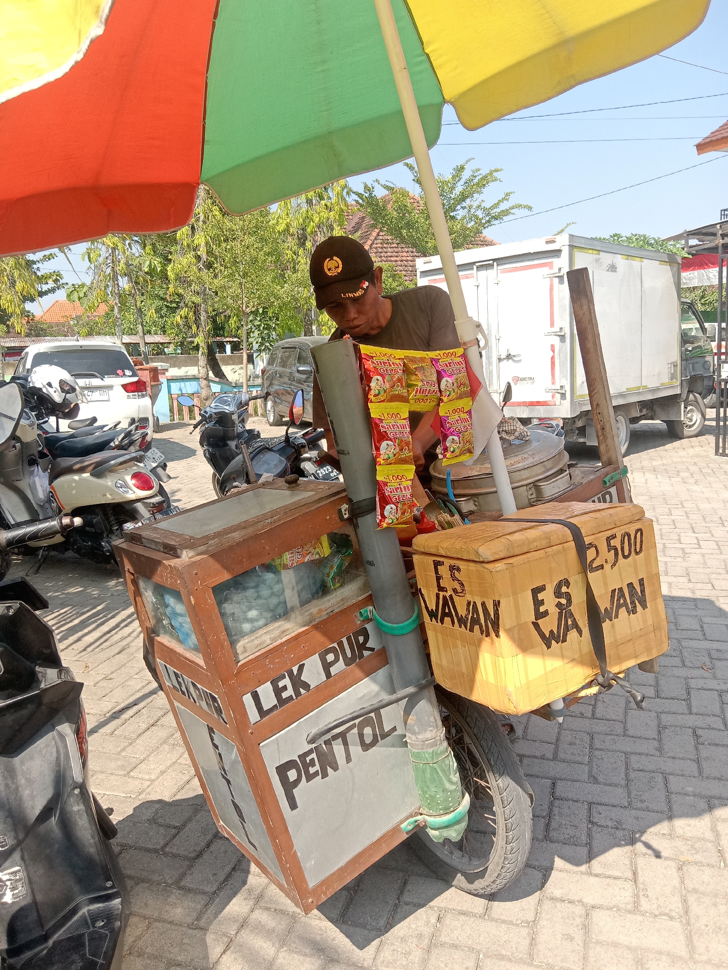 A man wearing a dark cap with a circular emblem stands behind a mobile cart on a paved street in Klepek, Indonesia. The cart is equipped with a large, colorful umbrella, red on the left and green and yellow on the right, providing shade. The man is focused on a collection of bagged snacks hanging from a pole attached to the cart. In front of him, a wooden cart with glass compartments displays various items, possibly food or drinks. A yellow cooler box with "ES WAWAN" and "2.500" written in black marker sits on the back of the cart. Several motorcycles are parked to the left and right of the cart. In the background, a white truck and other vehicles are visible, along with trees and buildings. The ground is paved with interlocking bricks, and shadows are cast by the umbrella and the cart.