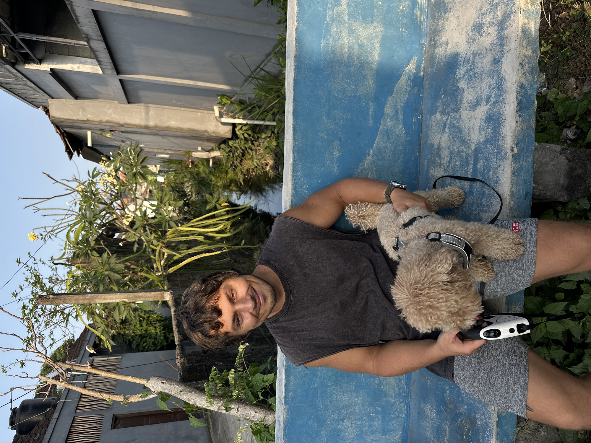 In the city of North Kuta, Indonesia, a man with brown, wavy hair and a beard is seated on a weathered blue bench. He is wearing a dark grey sleeveless t-shirt and grey shorts. He is holding a white and black dog leash in his left hand, and a light brown poodle is being held by his right hand. The poodle is wearing a harness. The man is smiling at the camera. In the background, there is a narrow waterway with lush green vegetation on either side, flanked by buildings. The sky is clear and blue.