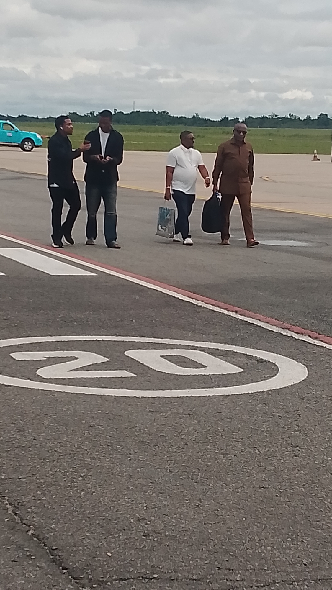 Four men are walking on an airport tarmac. The man on the far left is wearing a black jacket and black pants. Next to him, a man in a white shirt and jeans is looking at his phone. The third man from the left is wearing a white polo shirt and dark pants, carrying a blue and white patterned bag. The man on the far right is dressed in a brown suit and also carrying a dark bag. In the background, a turquoise vehicle is visible, along with a grassy field and trees under a cloudy sky. The ground has markings, including a white circle with the number "20" inside, indicating a speed limit. The scene appears to be during the daytime, with overcast weather.