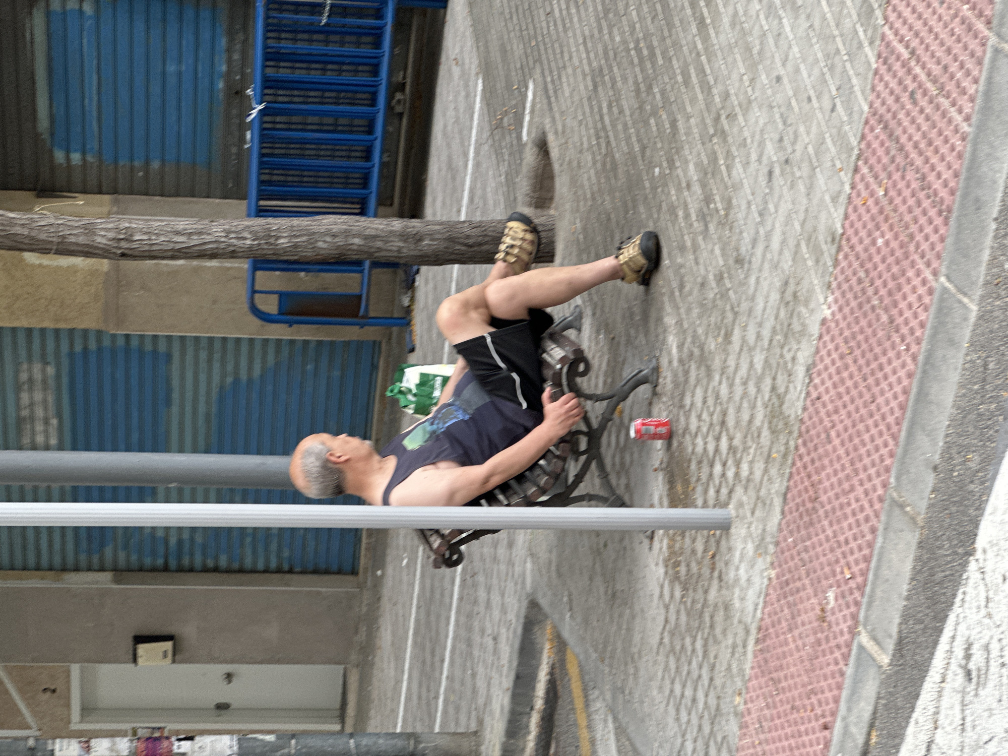 A man sits on a park bench in an urban environment, facing away from the camera. He is wearing a dark blue tank top and black shorts. His legs are crossed and he is wearing tan sandals. A red can sits on the ground near the bench. The bench is made of dark wood and metal. Behind the bench, a tree trunk rises vertically, and a blue metal fence runs horizontally. The ground is paved with grey and red patterned tiles. In the background, a building with a white door and blue shutters is visible. The scene appears to be a public sidewalk in a city.