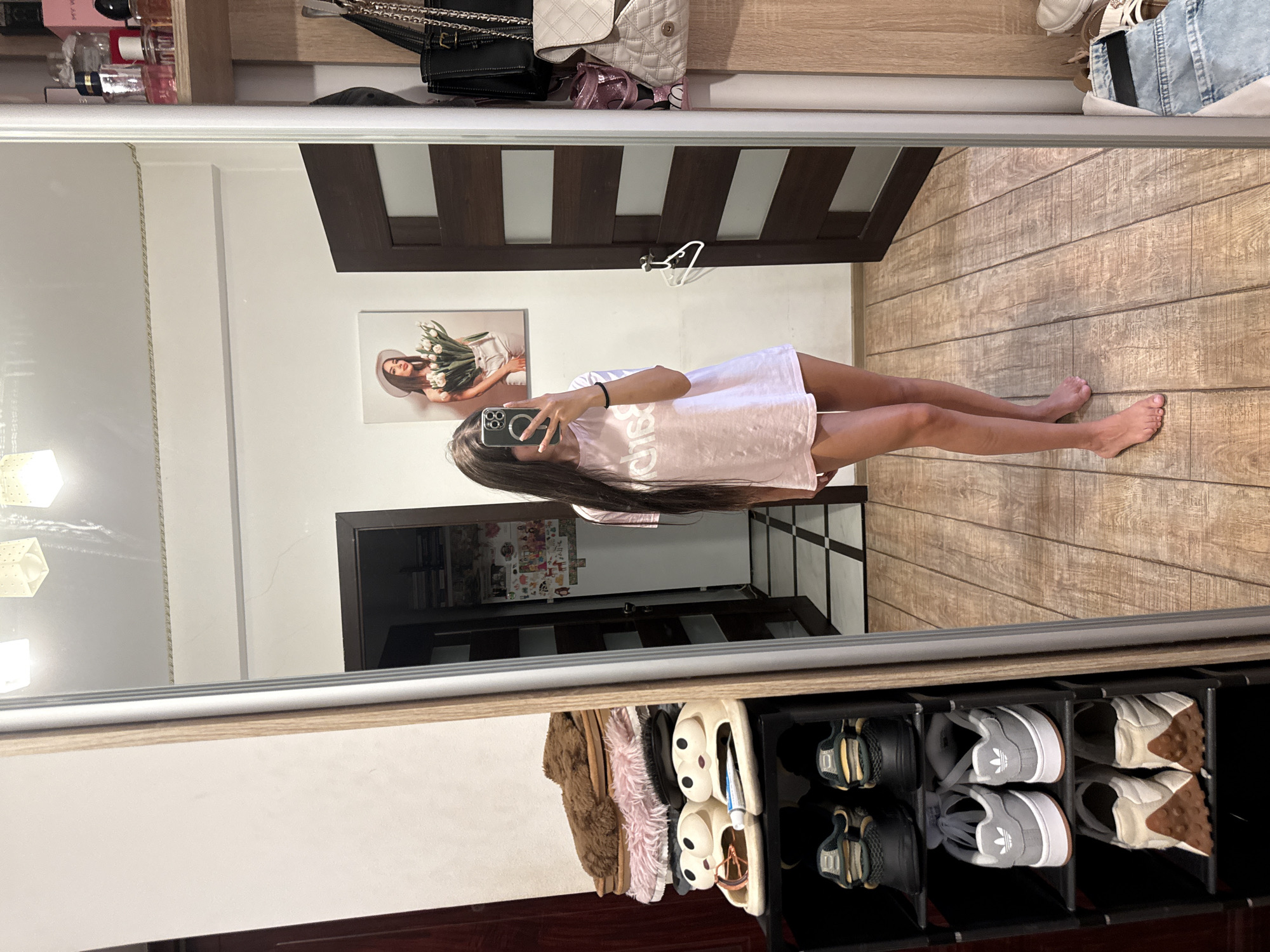 A young woman with long, dark hair stands in front of a full-length mirror, taking a selfie. She is wearing a light pink t-shirt and appears to be in a bedroom or dressing area. The mirror reflects a shoe rack filled with various shoes, a stack of slippers, a door, and a wall with framed pictures and shelves of toiletries. The floor is made of wooden planks. The image does not provide any specific indicators of its location in Chișinău, Moldova.