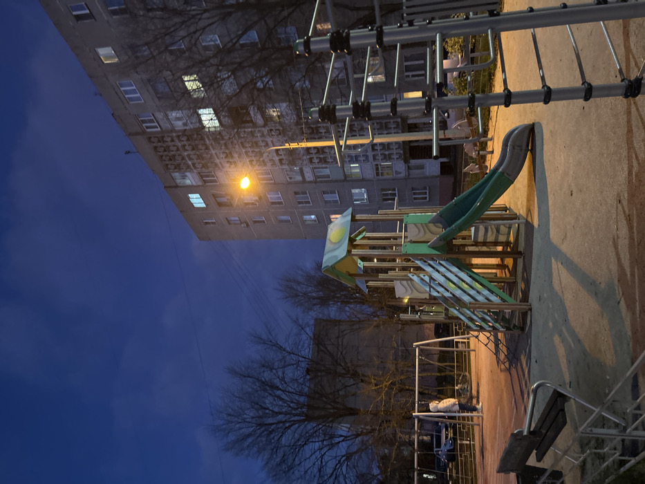 This image depicts a residential playground in Voskresensk, Russia, at twilight or night. A solitary individual, wearing a light-colored hooded coat, stands near a set of metal swings on the left side of the dirt-surfaced playground. In the center, a multi-component playground structure with a light green and yellow roof, wooden supports, stairs, a climbing net, and a dark green slide is present. To the right, a metal climbing frame with grey vertical posts and black horizontal bars is partially visible. A bright orange-yellow streetlamp, positioned in front of a multi-story grey residential building, illuminates the scene, casting long shadows across the playground. The building, prominent on the right, features numerous illuminated windows and a patterned facade. Bare deciduous trees are visible in the left background. The sky is a deep, dark blue with visible cloud formations.