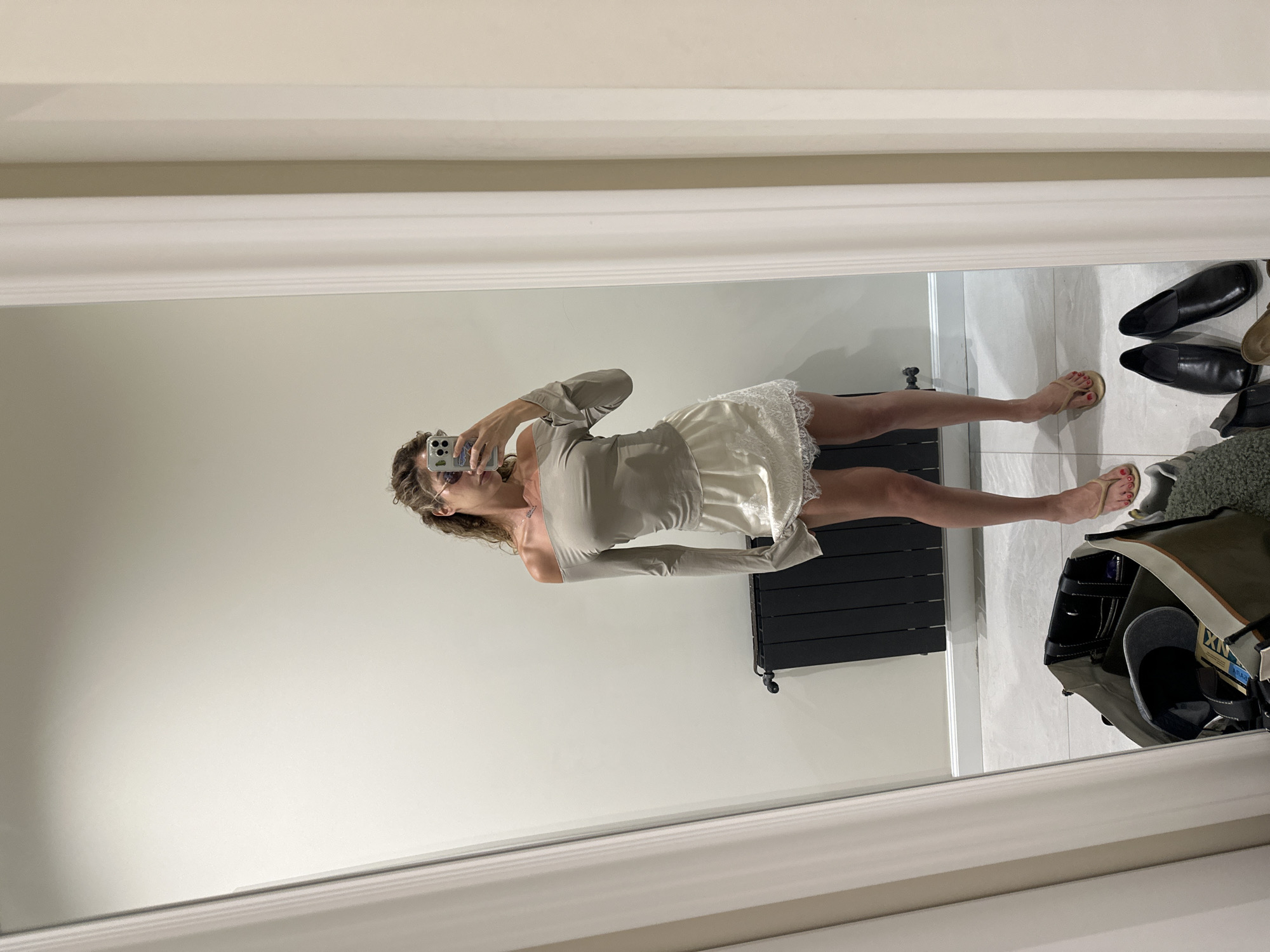 A woman stands in front of a large mirror, taking a selfie. She is wearing an off-the-shoulder taupe top and a white lace slip skirt. Her hair is curly and she is wearing sunglasses. Her feet are clad in beige flip-flops. In the reflection of the mirror, a black radiator is visible behind her, and to the left, a large canvas bag filled with various items, including a baseball cap and a wallet, is on the floor. Several pairs of shoes are also scattered on the floor in front of the mirror. The setting appears to be an indoor space, possibly a home, with neutral-colored walls and a tiled floor. The overall lighting is soft and appears to be from an overhead source. There is no indication that this scene is located in Tbilisi, Georgia.