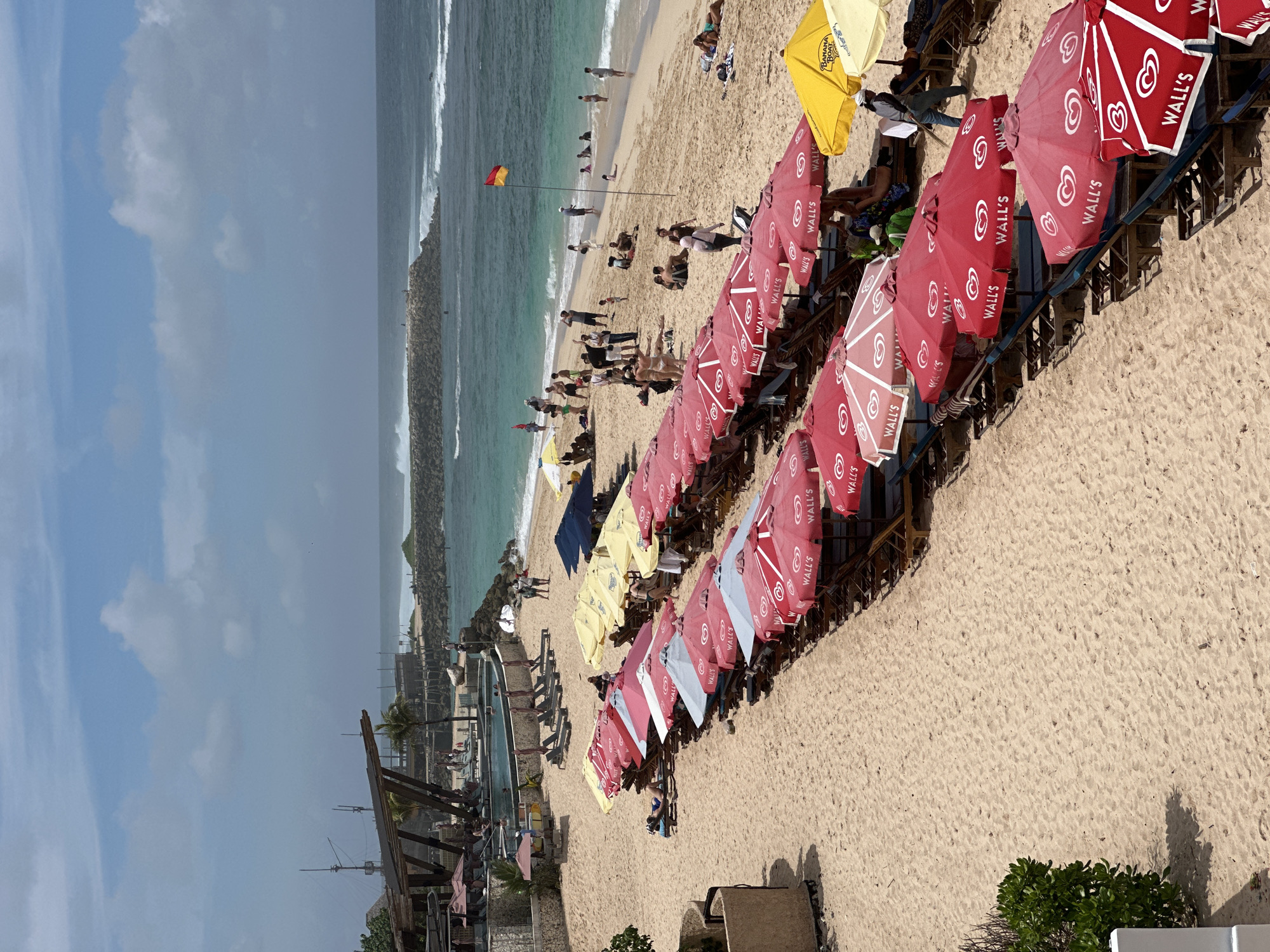 This vibrant image captures a bustling beach scene in Kuta Selatan, Indonesia, from an elevated perspective. The sandy shore is lined with numerous sun loungers and beach umbrellas, primarily bright red with "WALL'S" and a heart-shaped logo displayed, along with some yellow (one featuring "BANANA BOAT" text) and a few light blue/white umbrellas.
Many people are visible enjoying the sunny day: some are relaxing on the loungers, others are strolling along the shoreline, and several individuals are wading or standing in the clear turquoise ocean water. In the mid-ground, a yellow and red flag on a pole stands near the water, possibly marking a swimming area. The ocean exhibits moderate waves breaking along the shore, and a long rock barrier or breakwater extends into the sea on the left. Further down the beach to the right, a large swimming pool and a wooden structure, likely part of a resort or beach club, are visible. The sky is partly cloudy, suggesting good weather. The overall impression is one of a lively and relaxed tropical vacation spot during the daytime.