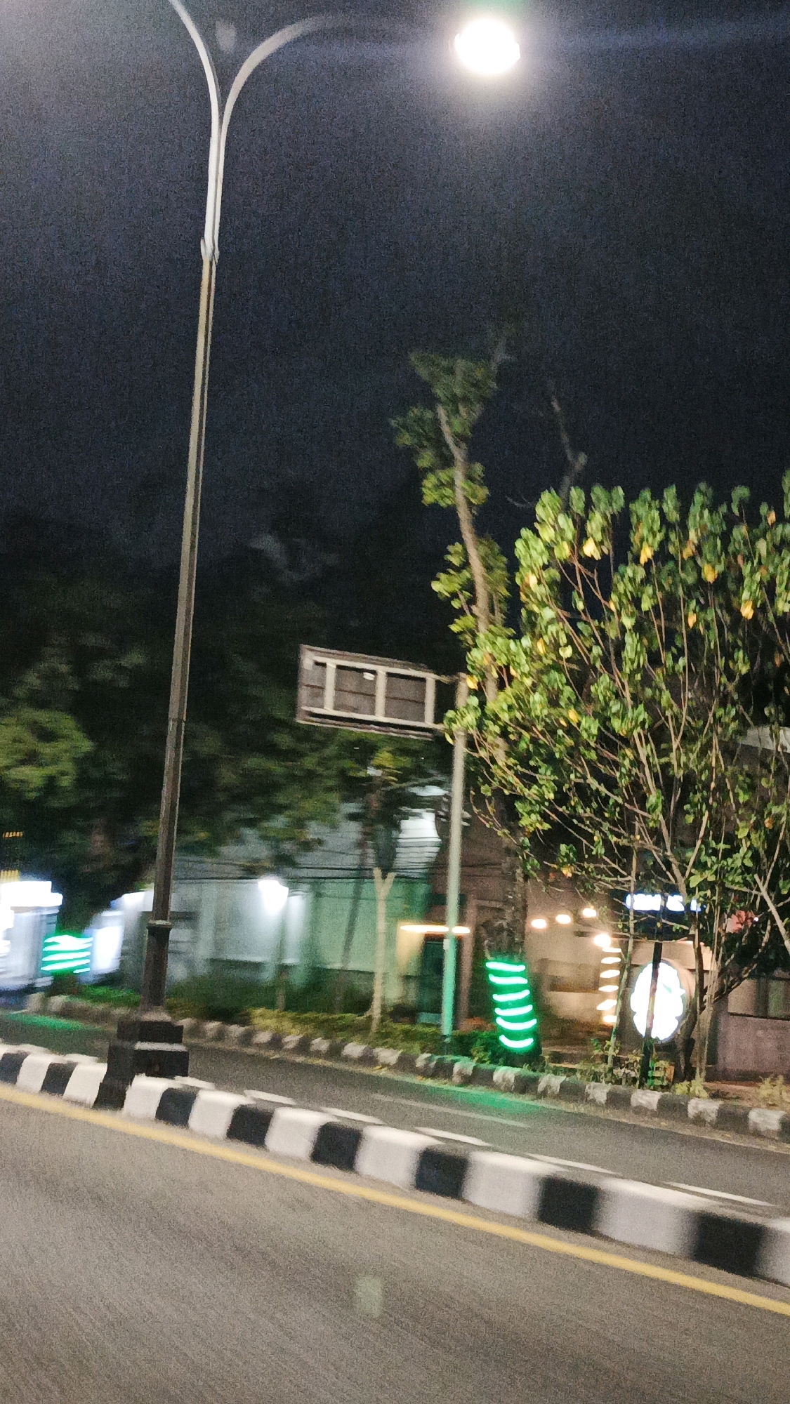 The image depicts a nighttime street scene in Surakarta, Indonesia. A tall, modern street lamp with a bright light illuminates the dark sky, with its pole bisecting the left side of the frame. To the right, a leafy tree with yellowish-green foliage is lit by the street lamp. In the background, a building with windows and illuminated signs is visible, along with some smaller trees and shrubs. The foreground features a paved road with a yellow center line and a curb marked with black and white stripes. Several neon green lights are present along the side of the road, some wrapped around tree trunks and others forming a curved shape. The overall impression is an urban environment at night, possibly near a commercial area or park, with decorative lighting.