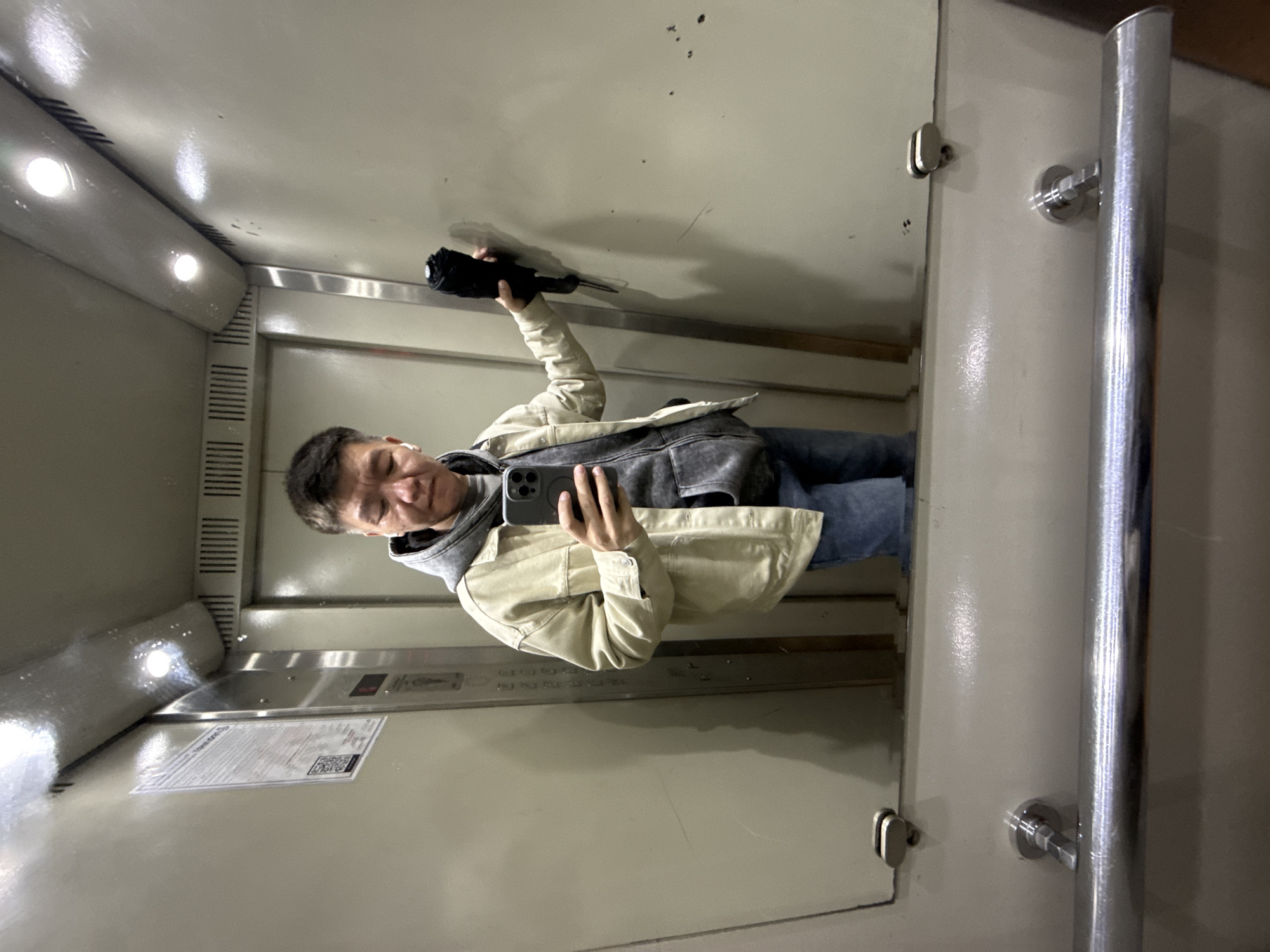 A young East Asian man is taking a mirror selfie in an elevator. He is wearing a light beige jacket over a grey hooded sweatshirt and blue jeans. He is holding a closed black umbrella in his right hand and a smartphone in his left hand, which he is using to take the photo. He has short, dark hair and is wearing white wireless earbuds. The elevator is clean and brightly lit, with metal walls and a metal handrail visible. A poster is attached to the left wall of the elevator. The elevator appears to be in a residential building, possibly in Khimki, Russia, given the context provided.