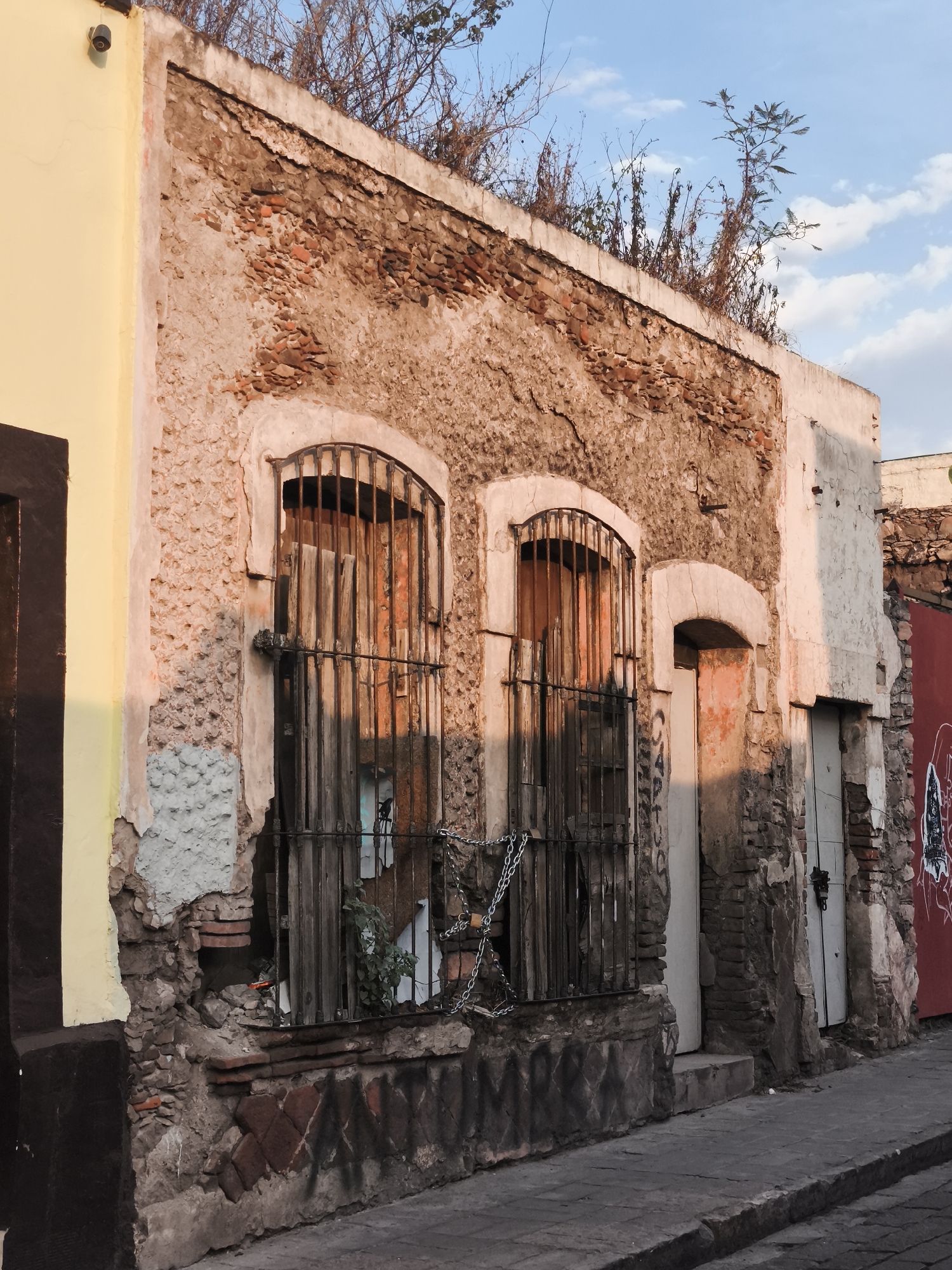 The image shows a dilapidated building in the city of Querétaro, Mexico. The building's facade is constructed of exposed brick and plaster, with two arched windows covered by iron bars and secured with a chain. The windows are boarded up with wood. To the right of the windows is a narrow doorway with a white door. Further to the right is another door, painted white, next to a red wall. The building appears to be in disrepair, with crumbling plaster and exposed brick. Plants and debris are growing on the roof of the building. The sidewalk in front of the building is cracked and uneven. The adjacent building on the left is painted yellow with a brown trim. Graffiti is visible on the walls. The sky is partially cloudy.