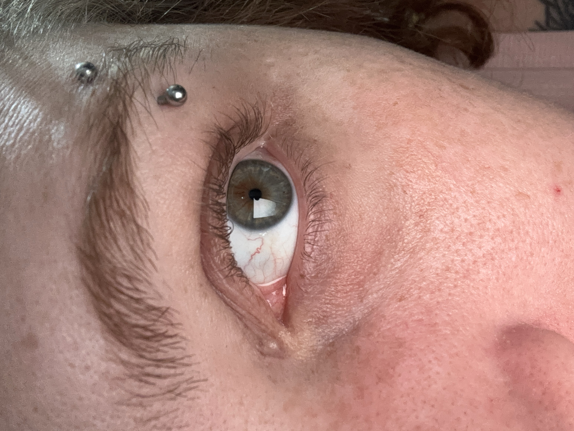 A close-up, angled view of a person's eye and surrounding facial features. The eye is light green with visible blood vessels in the sclera. The eyebrow is dark and thick with a piercing on the brow above the eye. The skin texture shows pores and a few blemishes. A small portion of the nose is visible in the bottom right corner of the frame. The location is not discernable.