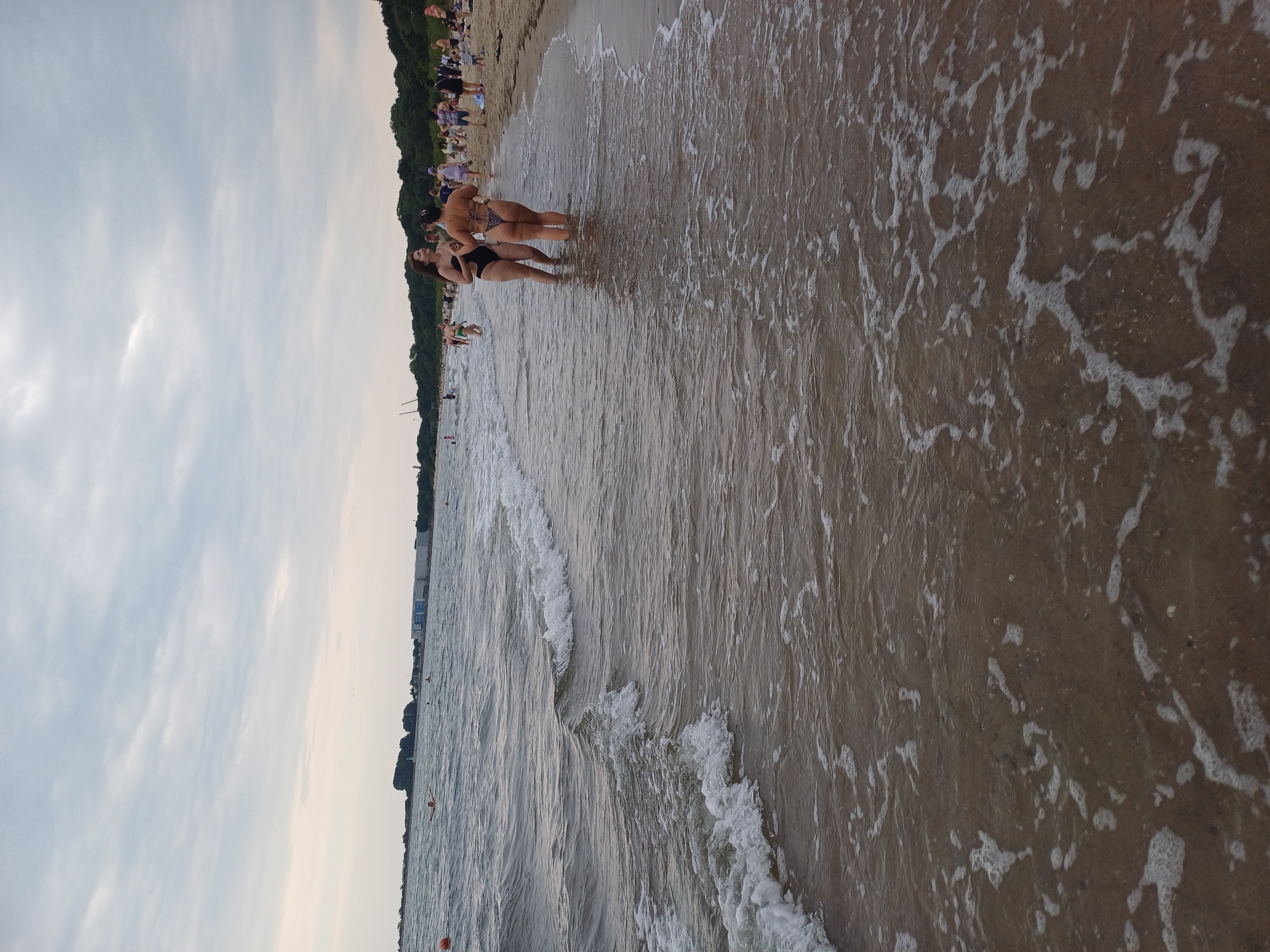 Two women in bikinis stand in shallow water on a beach, facing each other and talking. The woman on the left wears a black bikini, and the woman on the right wears a patterned bikini. Behind them, a crowd of people is visible on the sand, some walking, some sitting, and some in the water. The beach appears to be a public recreational area. The water is calm with small waves lapping the shore, and the sky is overcast. The background shows a treeline and some buildings, suggesting an urban or semi-urban environment. This scene is likely at the Gdańsk Beach in the city of Gdańsk, Poland, a popular destination for locals and tourists during the summer months.