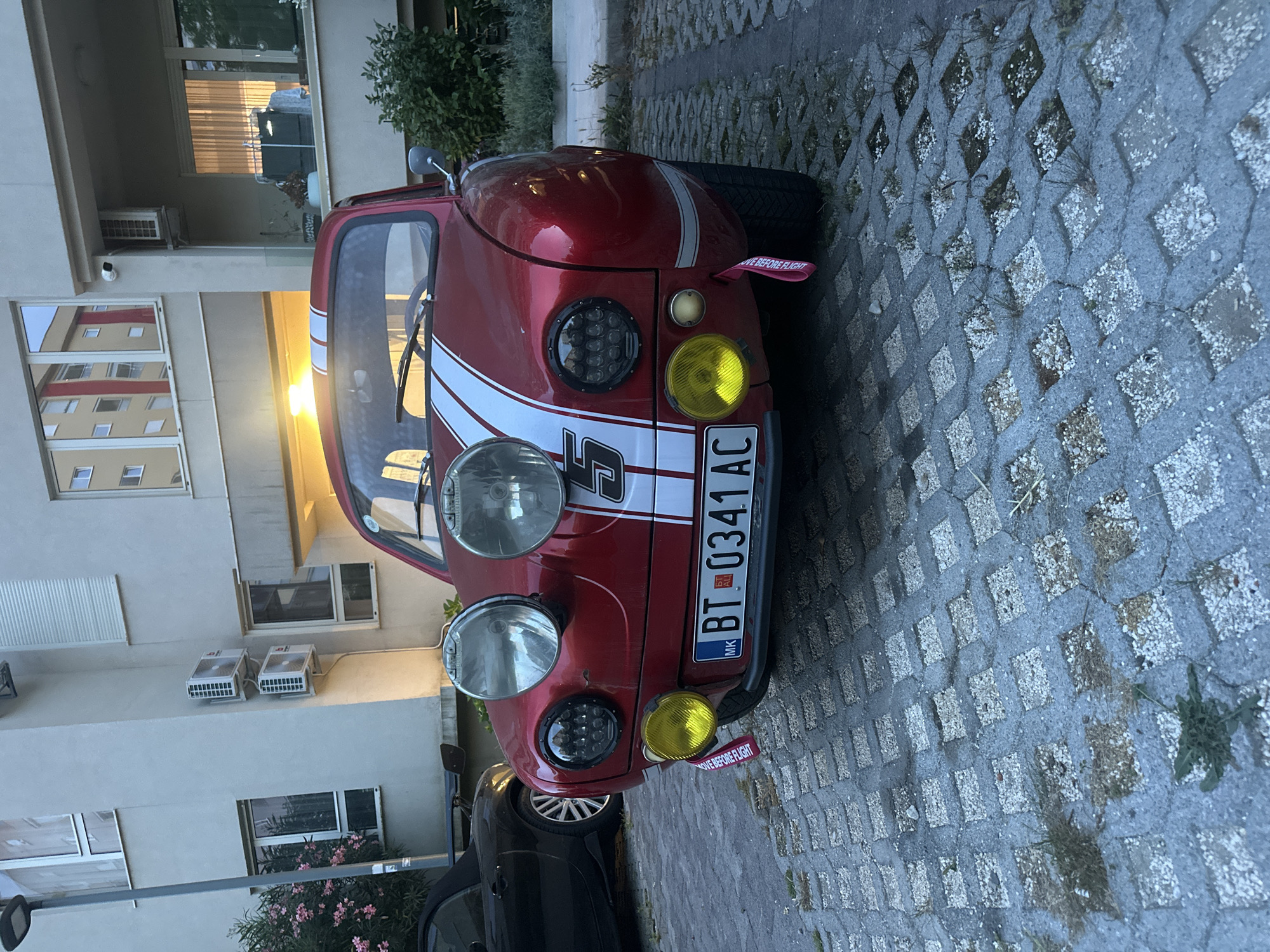 A red, custom-styled compact car, reminiscent of a classic Fiat 500 or similar model, is parked on patterned paver blocks next to a light-colored building. The car is adorned with prominent white racing stripes running over its hood and roof, featuring a large number "5" on the front fender. Its front fascia is distinctive, with two large, classic-style circular headlights, two smaller modern LED cluster headlights, and two yellow-tinted fog lights positioned below the main lights. A red "REMOVE BEFORE FLIGHT" tag hangs from the front bumper. The license plate, "BT 0341 AC", clearly shows "MK" (indicating North Macedonia) and "AL" (possibly referring to Albania or a specific region code) below the BT code, suggesting the vehicle is registered in Bitola, North Macedonia, even though the location context provided is Boreti, Montenegro.
The ground is composed of interlocking concrete pavers with diamond-shaped openings, many of which contain gravel, pebbles, and sparse grass or weeds. In the background, a multi-story building features several windows reflecting the surroundings, and two exterior air conditioning units are mounted on its facade. A security camera is also visible on the building. The lighting suggests late afternoon or early morning, with a warm glow from the sun reflecting off the car's surface and parts of the building. No people or specific activities are visible; the scene depicts a static, outdoor parking environment.