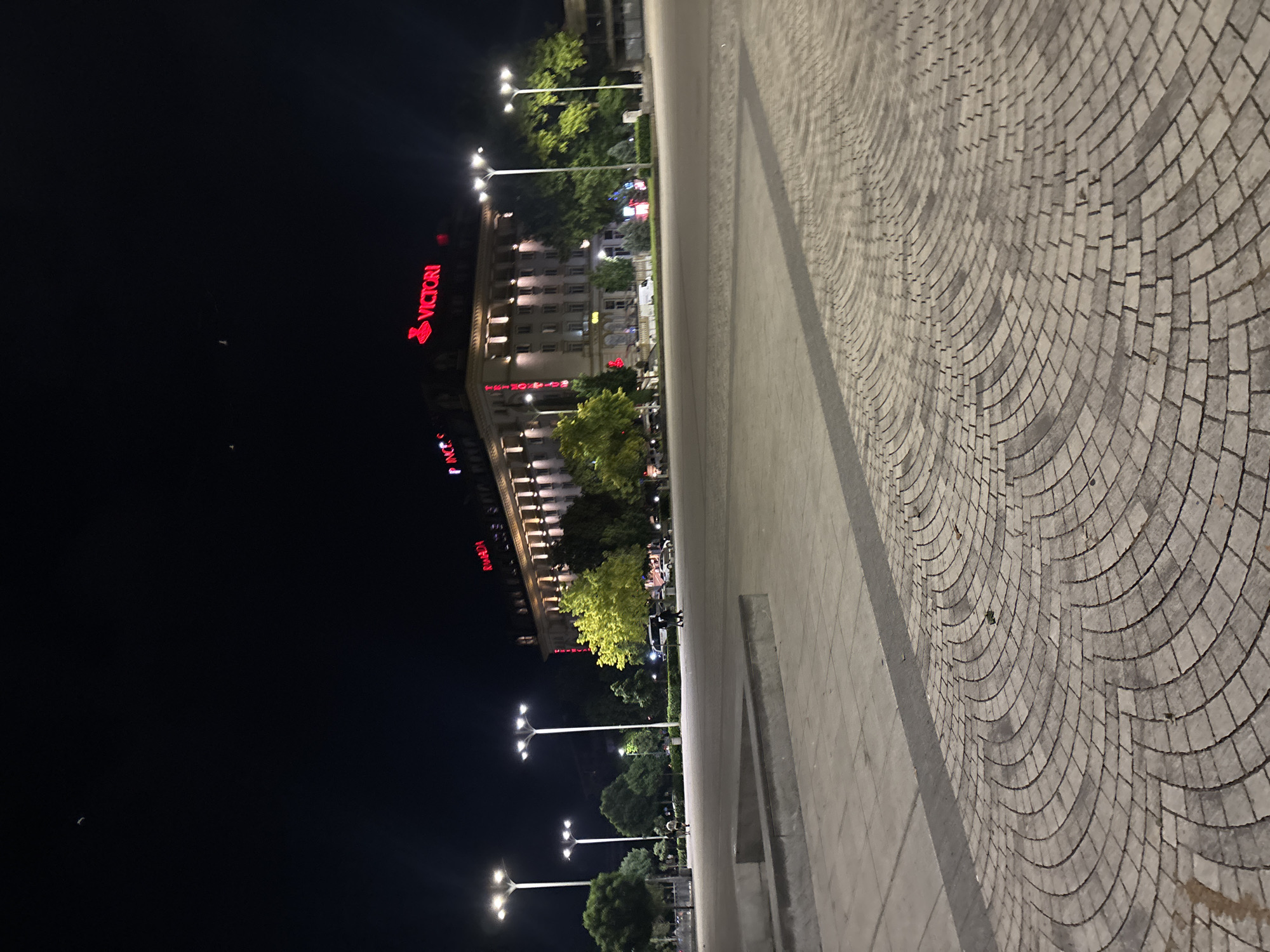 The image depicts Vazrazhdane Square in Sofia, Bulgaria, at night. The background is dominated by the Ramada by Wyndham Sofia City Center hotel, a multi-story building with a light-colored facade featuring numerous windows, columns, and cornices. Red neon signs on the building's roofline display "VICTORI" (likely part of "VICTORIA") centrally and "RAMADA" on the right side. A partial red neon sign, possibly "PINC S," is visible on the far left of the building. The hotel's lower floors are illuminated by upward-facing floodlights.
The vast foreground and midground consist of a paved square. The immediate foreground features a light-grey paver pattern arranged in concentric arcs. A darker straight line of paving separates this area from a uniformly paved section further back. A low, rectangular concrete or stone structure is positioned in the left foreground.
The square is brightly lit by at least five tall, modern streetlights, each with multiple cool-white LED fixtures. Various trees with green foliage are planted around the perimeter and within the square, with some appearing particularly vibrant due to artificial illumination. In the midground, a few individuals are sparsely distributed: two people walk together towards the right near a brightly lit green tree, and one person walks towards the left further back. Several vehicles, including a white van and other cars, are visible on the street adjacent to the hotel.
The sky is dark, indicating nighttime. A faint crescent moon is discernible in the upper left quadrant. Several small, indistinct white objects are present in the upper sky above the hotel building.