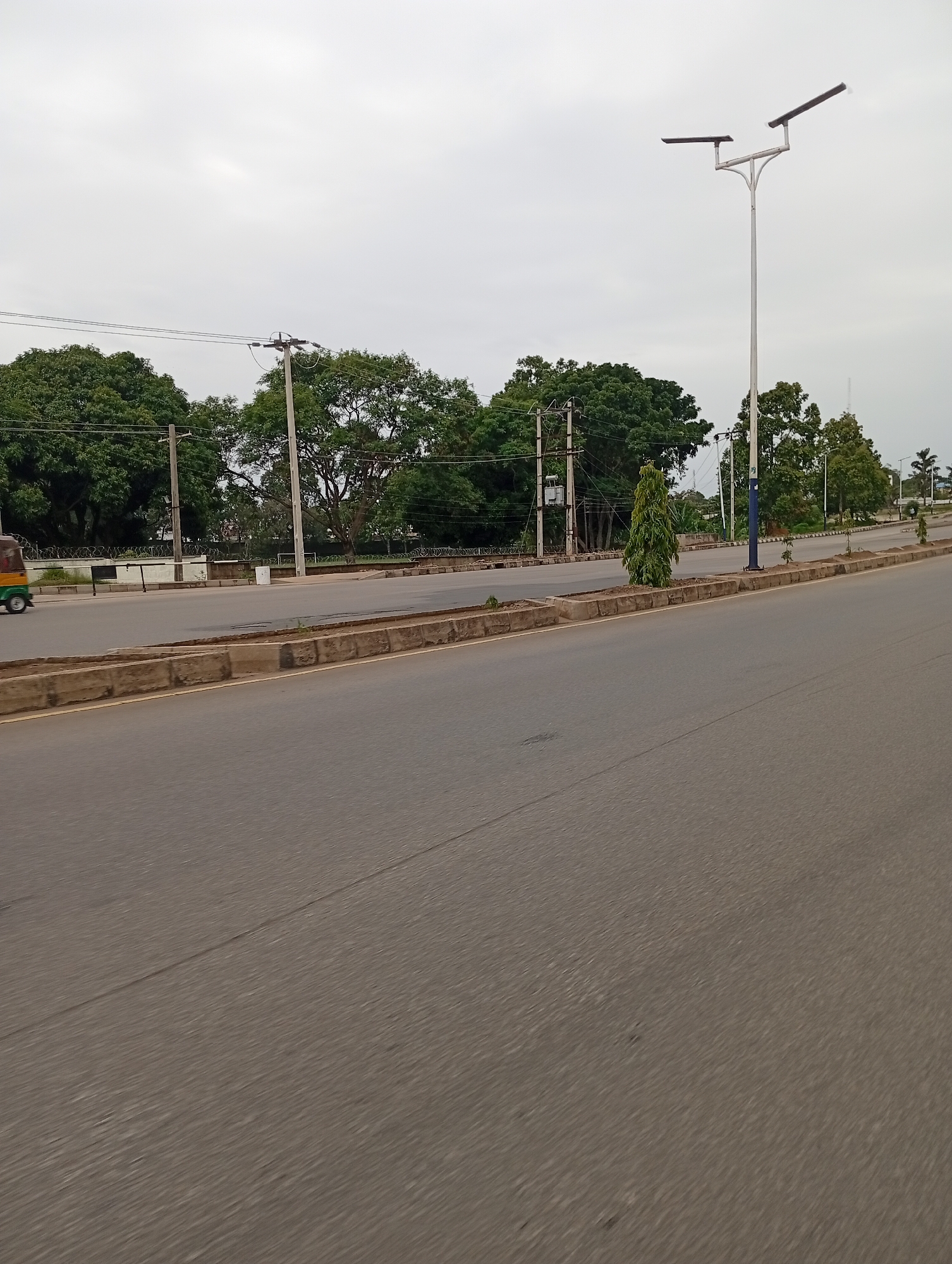The image shows a street scene in Jos, Nigeria, with a paved road in the foreground. A median strip separates the road from a sidewalk and a line of trees. A traffic light pole with two lights is visible on the right side of the image. In the background, there are more trees and buildings, as well as some power lines and poles. An auto-rickshaw is visible on the left side of the road. The sky is overcast.