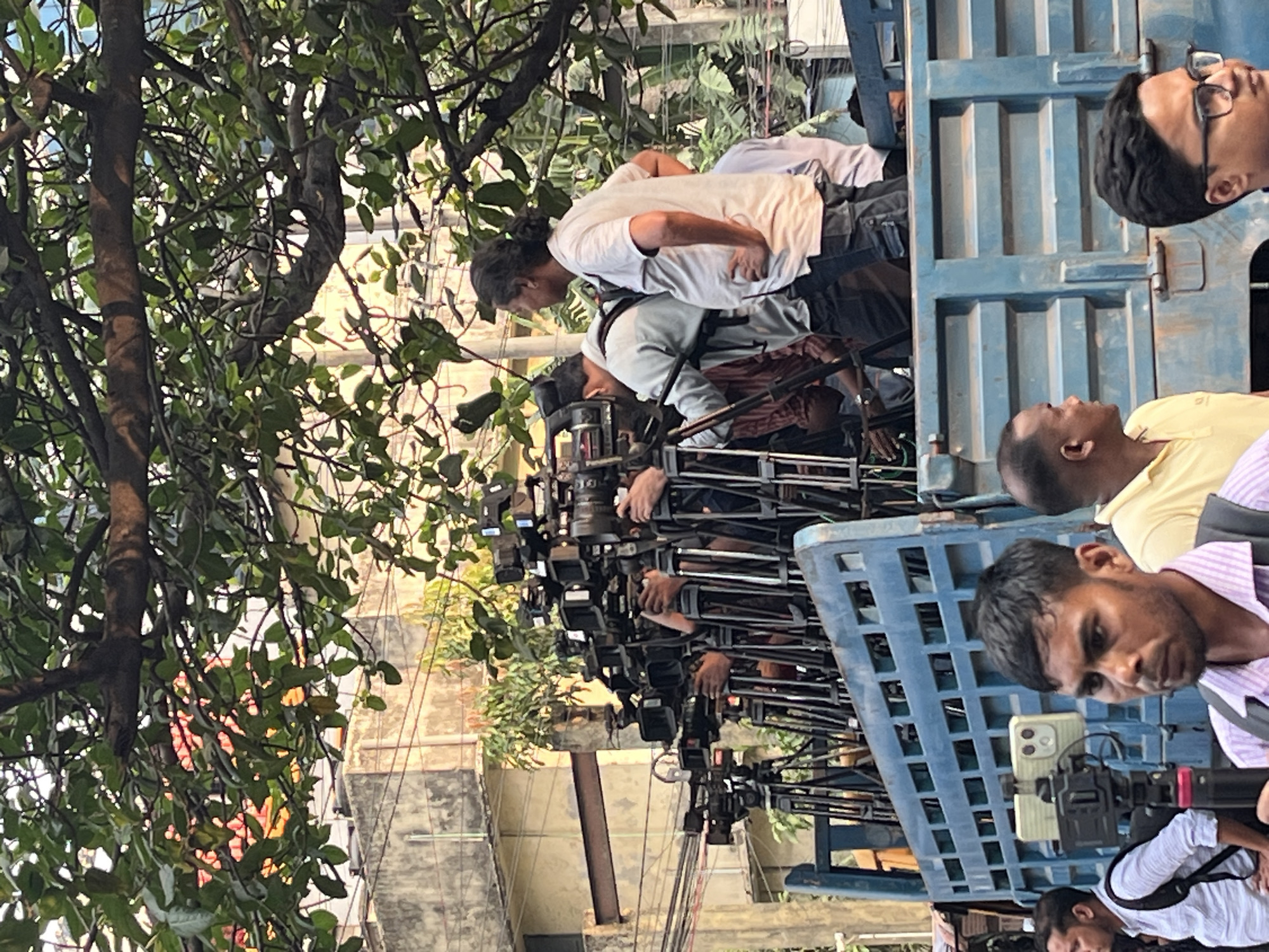 A group of people are standing on the back of a blue flatbed truck, holding cameras on tripods. The people on the truck seem to be filming something, and there is a crowd of cameras. The truck is parked on a city street in Dhaka, Bangladesh, as evidenced by the building style, foliage, and the appearance of the people. The scene is busy and appears to be a news event or a public gathering being documented.