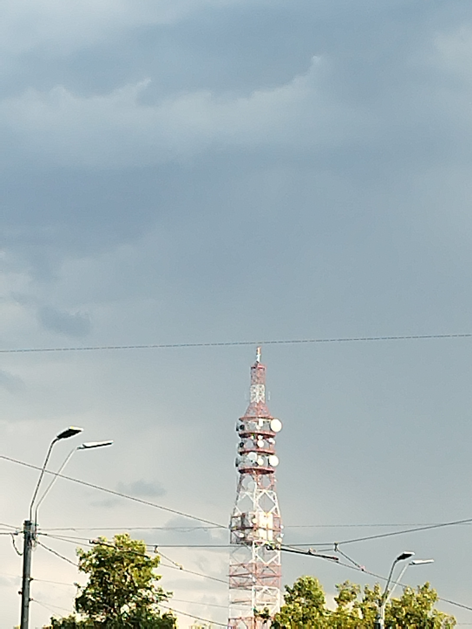The image shows a tall, lattice transmission tower with a white and red exterior, located in Bucharest, Romania. The tower is adorned with numerous circular antennae at various levels. In the foreground, a street lamp with two lights is visible on the left side of the frame, and a section of lush green trees is present at the bottom. Overhead power lines crisscross the scene, and the sky is overcast with gray clouds. The tower is a prominent structure in the urban landscape, suggesting its function as a telecommunications or broadcasting hub. The overall atmosphere is one of a typical city environment under a cloudy sky.