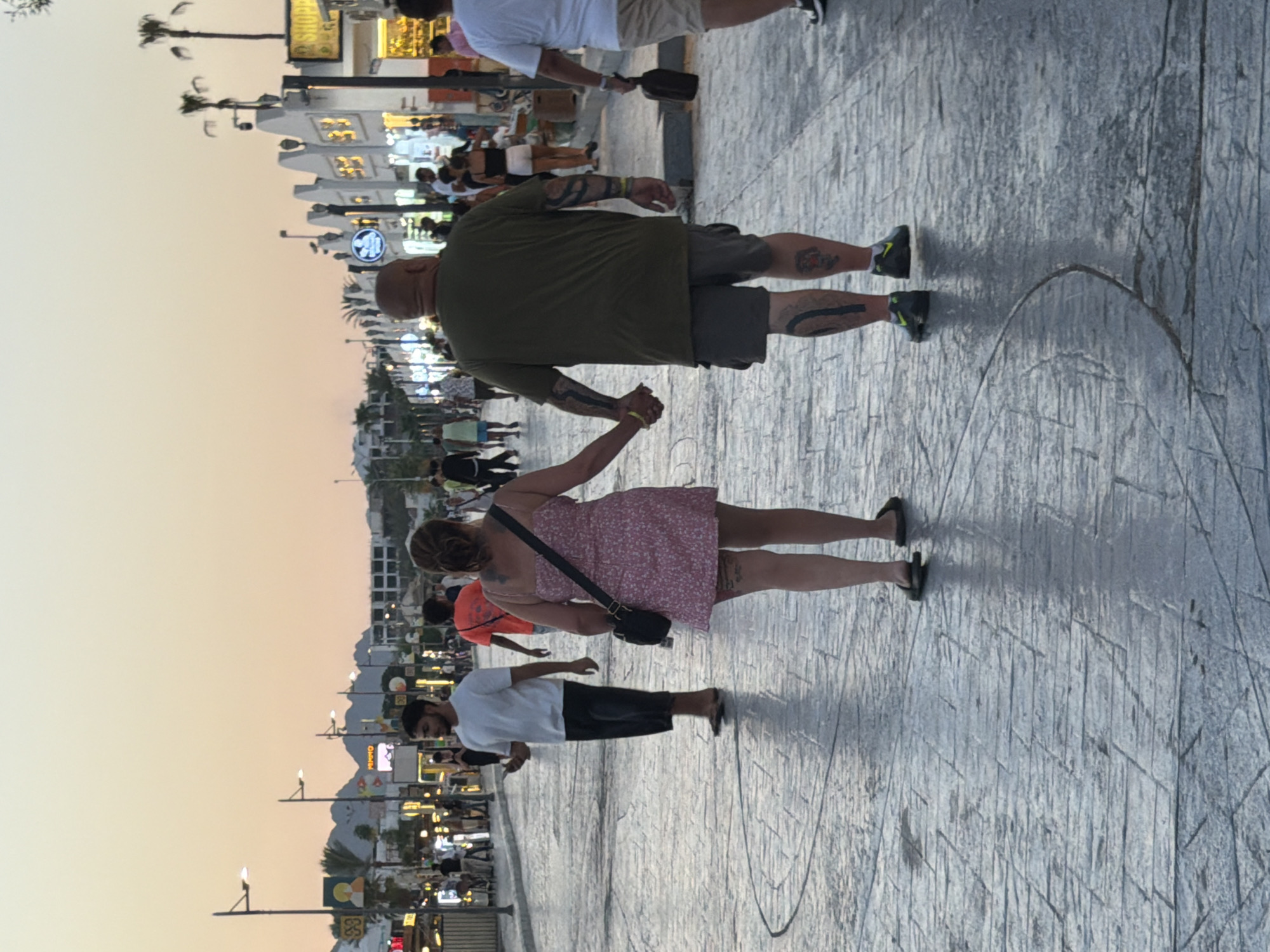 The image depicts a scene on a street in Sharm El Sheikh, Egypt, during the late afternoon or early evening, as suggested by the warm, diffused light and the pale orange sky. The street is paved with large, textured rectangular stones that appear slightly damp, reflecting the ambient light and creating a patterned surface.
In the foreground, a couple is walking away from the camera, holding hands. The woman on the left wears a pink floral print dress and carries a black shoulder bag. She has tattoos visible on her legs. The man on the right wears a dark green t-shirt and dark grey shorts. He has numerous tattoos on his arms and legs. He is wearing dark sneakers with yellow accents.
Behind them, other pedestrians are visible walking in both directions. To the left, a man in a white t-shirt and black shorts is walking towards the camera, holding a smartphone. Further back, more people and a row of shops with illuminated signs line the street. Palm trees and buildings are visible in the background, with hills or mountains in the distance under the dusky sky. The shops have a distinct architectural style with pointed, angular roofs and decorative elements. The overall atmosphere is that of a busy pedestrian walkway in a tourist area.
