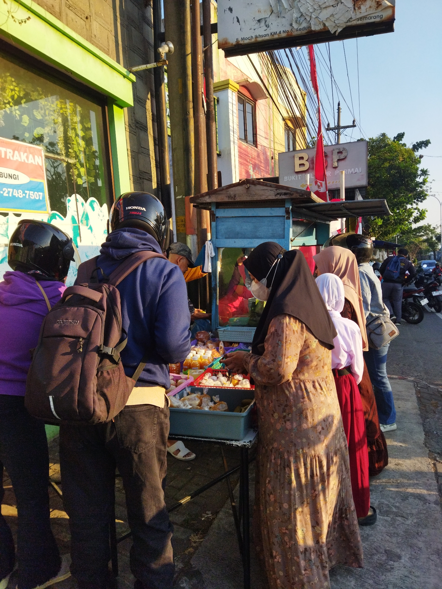 The image captures a street scene in Semarang, Indonesia, with a focus on a food stall situated alongside a road. Several individuals are gathered around the stall, some appearing to be customers and others possibly vendors. The atmosphere suggests a bustling moment, possibly during the day given the sunlight casting shadows.
In the foreground, two individuals in motorcycle helmets are visible from behind, one wearing a purple hoodie and the other a blue hoodie with a backpack. They are facing the food stall. Further down the line of people, a woman wearing a hijab and a floral dress, with a face mask on, is interacting with the stall. Other people, including a child in a white head covering and a red skirt, are also present, suggesting a social gathering or a popular spot for snacks.
The food stall itself is a small, open-air structure with a blue awning. It displays various food items, including what appear to be packaged snacks and bottled drinks. Behind the stall, a building with yellow and pink paint can be seen, along with signs, one of which indicates "BUKIT INDAH PERMAI". Electrical wires are strung overhead, a common sight in urban Indonesian settings. In the background, motorcycles are parked and a few vehicles are on the road, indicating the location is on a public thoroughfare. The overall scene is one of everyday life unfolding on a city street.