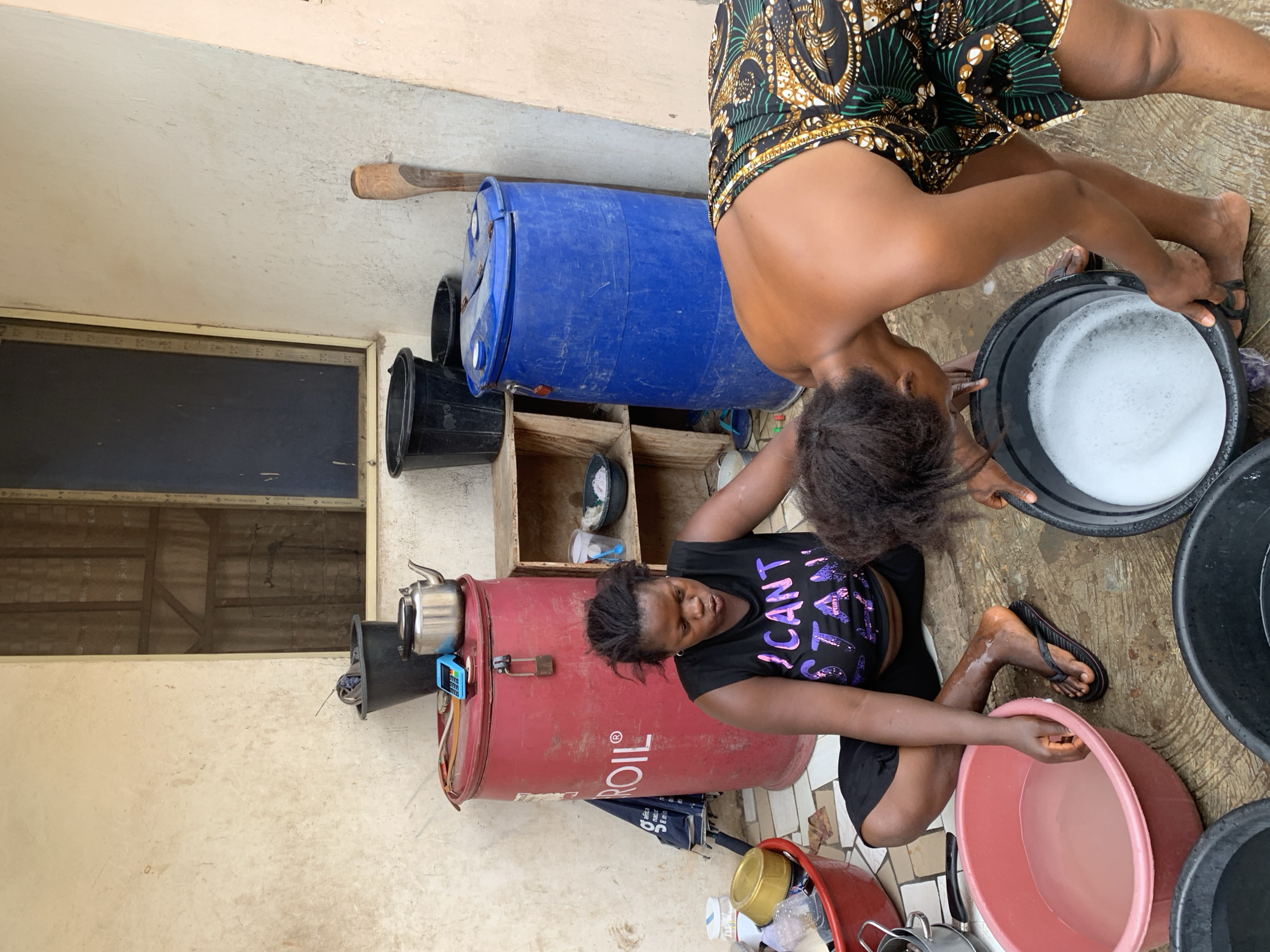 Two women are engaged in laundry activities outdoors in what appears to be a domestic space in Lokoja, Nigeria. One woman, wearing a printed top, bends over a black bucket filled with soapy water. The other woman, wearing a black t-shirt with a graphic and black shorts, kneels beside a red oil drum and several buckets of water. There is a window in the background of a white wall, alongside buckets, storage containers, and household items scattered around. A blue water barrel is behind the kneeling woman.