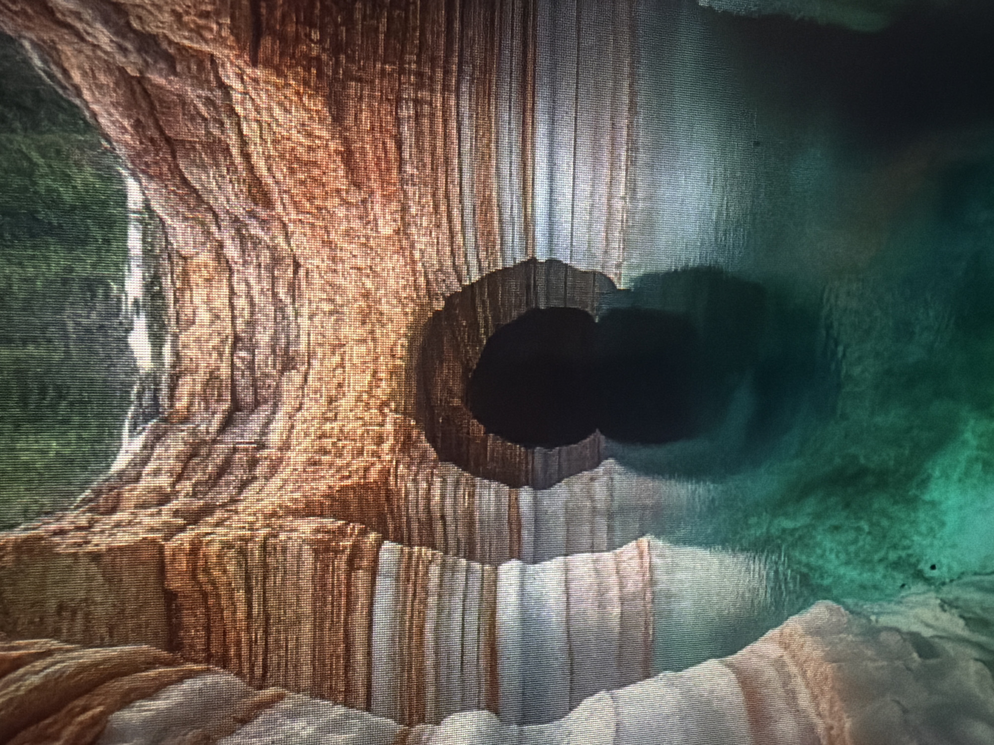 The image depicts a scenic view of a cave entrance opening into a body of water. The cave walls are layered with natural rock formations, exhibiting various shades of brown, orange, and white. The water is a vibrant turquoise and appears to be still, with reflections of the cave walls visible on its surface. The entrance of the cave is a dark, shadowed opening. Above the cave, a blurred green area suggests foliage or trees. The image is presented with a noticeable digital artifacting or pixelation, particularly visible in the vertical lines across the entire frame. There are no people or animals visible in the scene. The location of Baan Mae Nam, Thailand, is not identifiable from the visual information provided in the image.