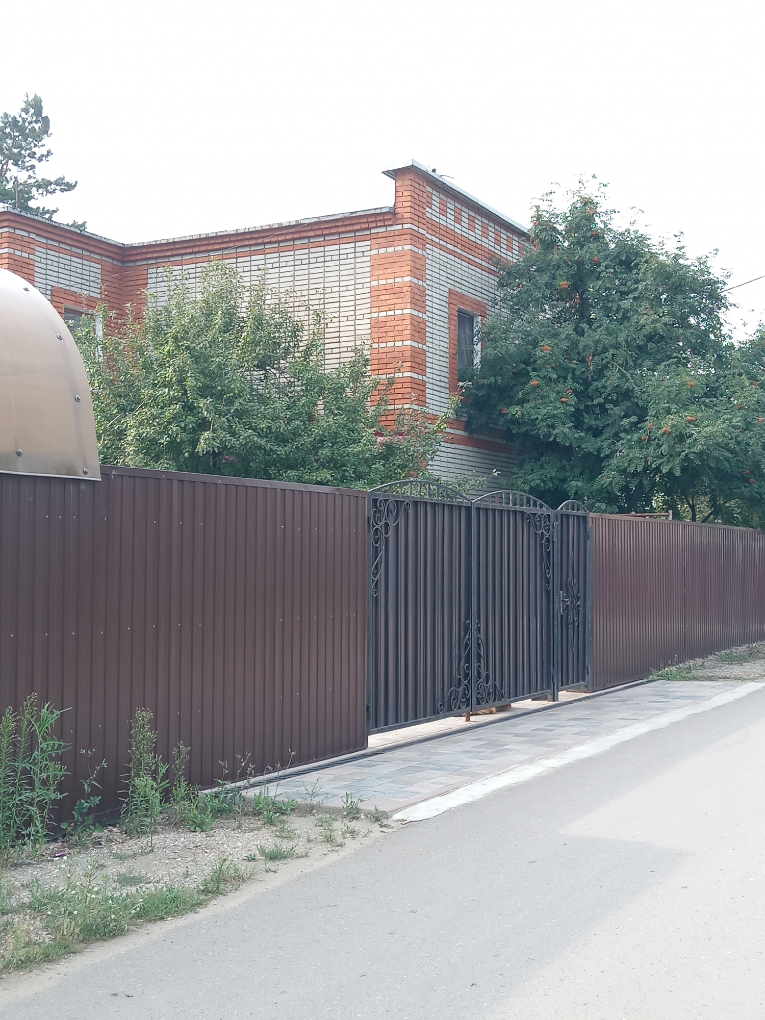 The image depicts a residential property in Rudny, Kazakhstan, characterized by a tall, dark brown corrugated metal fence. A section of the fence features an ornate, dark metal gate with decorative scrollwork. Behind the fence, a brick building with red and white accents is visible, surrounded by lush green trees, some of which bear clusters of red berries. To the left of the gate, a light-colored, arched structure, possibly a carport or shed roof, extends over the fence line. The foreground shows a paved walkway adjacent to the fence, bordering a grey asphalt road. The area between the fence and the road is sparsely vegetated with weeds and small plants, and scattered gravel. The sky is overcast and bright.