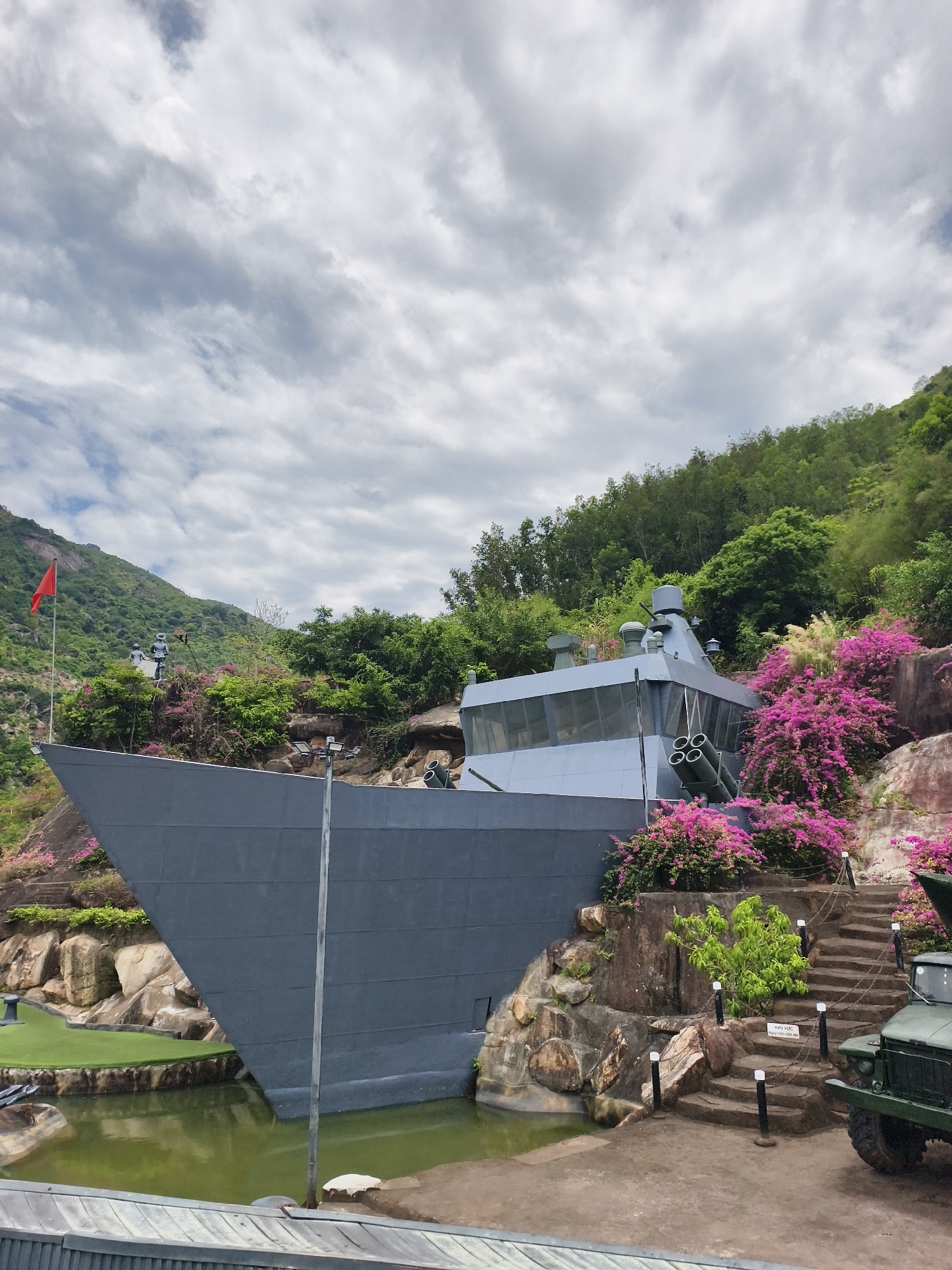The image shows a gray ship structure located in Tay Nha Trang Ward, Vietnam, near a hillside densely covered in vegetation. The ship is grounded near a small pond. A red flag flies atop a hill behind the ship. Pink and green plants surround the ship structure. A set of stone stairs with chained posts leads up the hill near the ship. Part of a military vehicle is visible on the right. The sky is cloudy.
