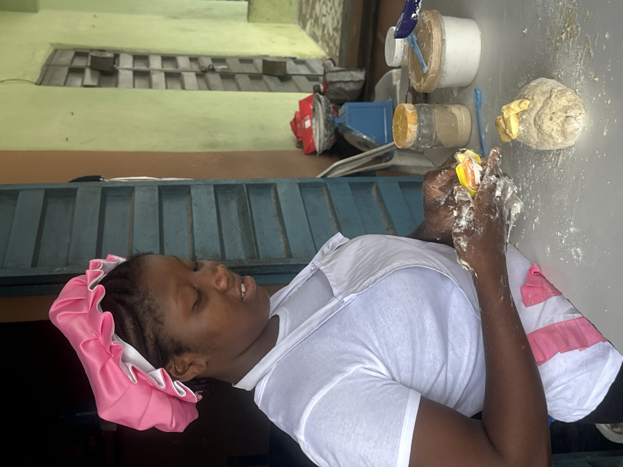 The image depicts a young Black woman wearing a pink bonnet and a white apron over a white t-shirt, engaged in food preparation. She is standing in what appears to be a workshop or kitchen environment, evidenced by the presence of various ingredients and cooking implements on a table. In the foreground, she is holding a small packet, possibly containing yeast or a similar baking ingredient, with her hands dusted with flour. A ball of dough with small pieces of what looks like butter or margarine on top sits on the table in front of her. Behind her, the background includes a blue-green door or window frame, an orange wall, and a light green wall, suggesting an indoor setting. Further in the background, a blue weighing scale with a red bag on top is visible, along with containers of flour and other ingredients. The overall scene suggests a baking activity taking place in a domestic or small commercial setting within Badagry, Nigeria.