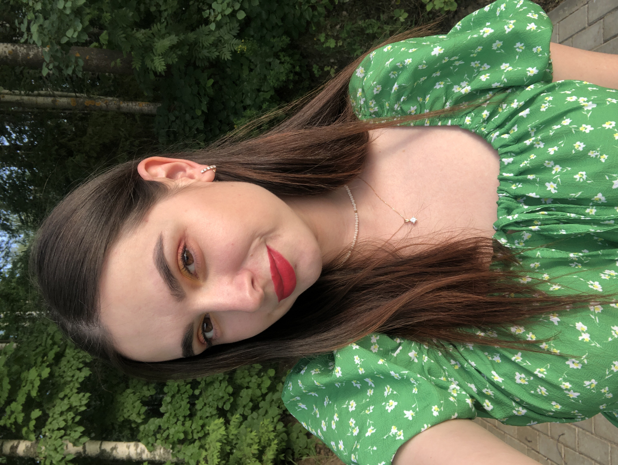 A young woman with long, brunette hair and red lipstick is wearing a green floral dress and posing for a photo outdoors. She is wearing a sparkly earring and a delicate necklace. The background is blurred green foliage, and there are trees in the distance.