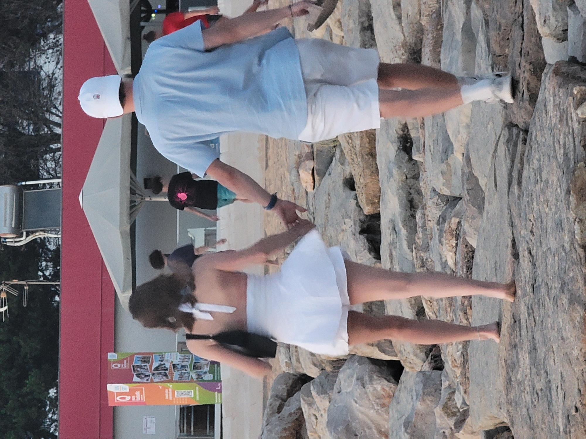 This media file depicts two people near a rocky shoreline, likely in Quarteira, Portugal. The primary subject is a person in the foreground wearing a white dress and a black purse. They are facing away from the camera and their back is visible. Their hair is dark and styled with a white ribbon. Behind them, a person in a light blue shirt and white shorts is seen from the waist up, also facing away from the camera. This individual is wearing a white baseball cap.
The scene appears to be outdoors, with a backdrop of large, rough-textured rocks. The lighting suggests it might be late afternoon or early evening, with a warm glow on the rocks. The overall atmosphere seems casual and relaxed, possibly a vacation or a leisurely outing.
The background is slightly out of focus, but a red structure is visible on the left, possibly a building or part of a beachside establishment. There are also some posters or signs visible on this structure, but any text is unreadable due to the image quality and distance. The presence of rocks and the open, outdoor setting are the most prominent cues to the location. There is no clear indication of specific weather conditions beyond what might be inferred from the lighting. The activity appears to be people enjoying the seaside environment.