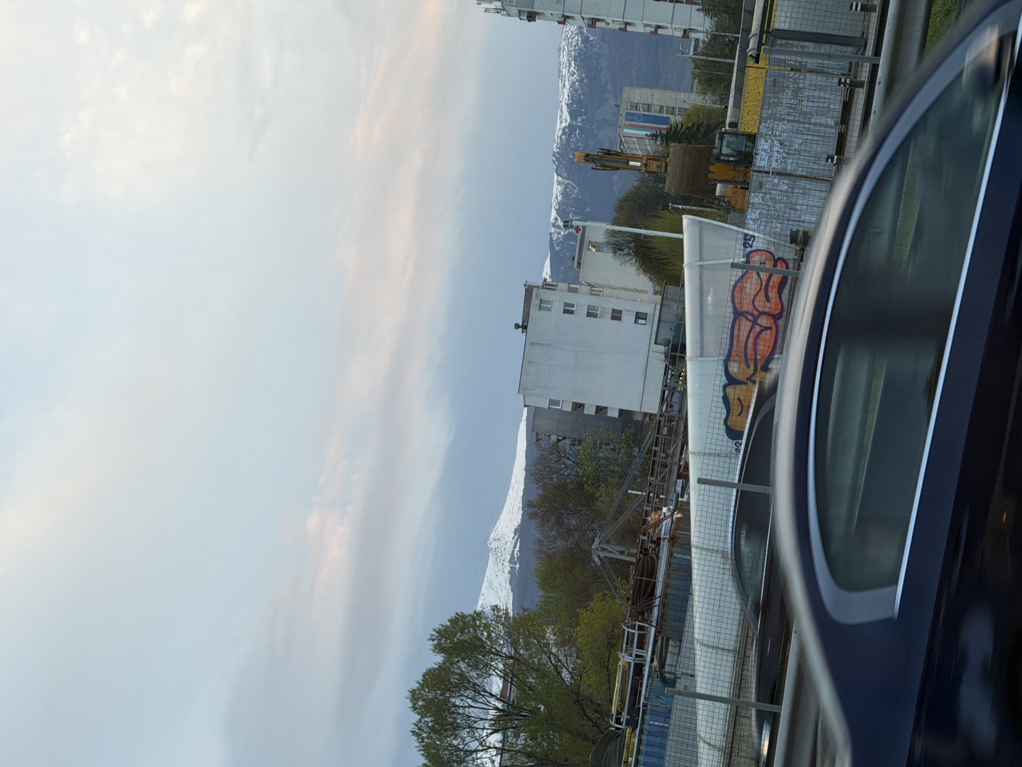 The image, captured from the interior of a vehicle, showcases an urban or semi-urban landscape with prominent snow-capped mountains in the background. In the immediate foreground, the side mirror and part of the dark-colored body of a car are visible on the right. To the left, a concrete barrier or wall is adorned with abstract, curvilinear graffiti in shades of orange and red.
The middle ground reveals an area that appears to be under construction or industrial, featuring various metal fences, scaffolding-like structures, and a distinct yellow excavator. Several green trees with full foliage are scattered throughout this section.
Further back, multi-story buildings, predominantly white or light-colored, line the scene. One building in the mid-left background displays a banner or design with horizontal stripes, possibly blue, white, and red, resembling a national flag. Beyond these structures, imposing mountains, their peaks covered in snow, stretch across the distant horizon under a soft, cloudy sky.
The lighting suggests either early morning or late afternoon, with diffused light filtering through the overcast sky. The snow on the mountains indicates a colder season, such as winter or early spring/late autumn. No discernible text is present beyond the abstract graffiti on the wall.