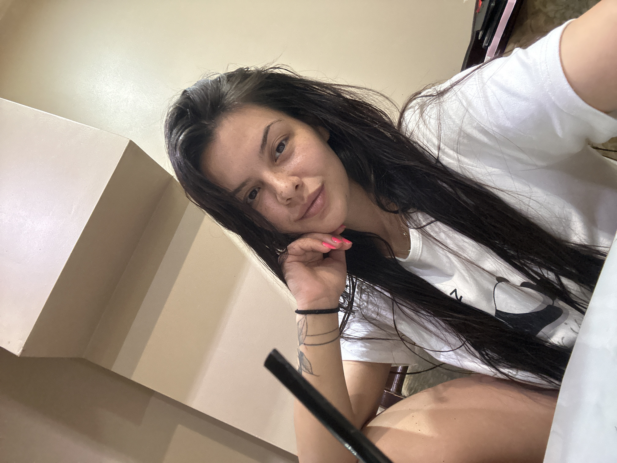 A young woman with long, dark hair is captured in a selfie taken indoors. She is wearing a white t-shirt and has a tattoo on her left arm. Her right hand is resting on her chin, and her nails are painted a bright pink. The background appears to be a room with light-colored walls and architectural features that suggest a modern interior. There is no indication of the city of Sliven, Bulgaria, or any other specific location within the image. The lighting is soft, and the focus is on the woman's face.