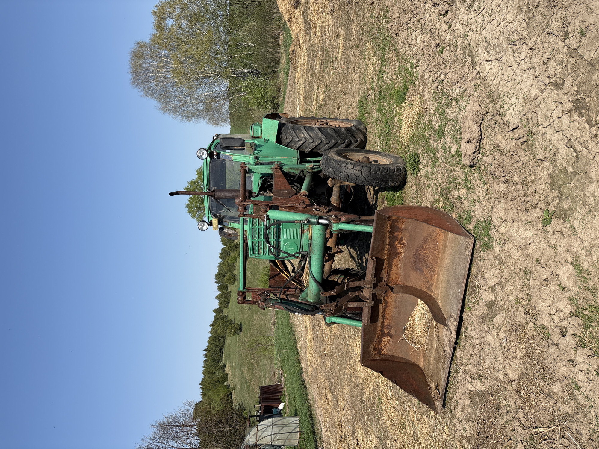 The image shows a green tractor with a rusty front loader parked on a dry, cracked field. The ground is sparsely covered with dead grass and small patches of green vegetation. In the background, there is a line of trees on a slight hill, and a clear blue sky above. In the distance to the left there is a small building or structure.
The image does not contain enough information to confirm the location of this scene in the city of Lukintsy, Russia.