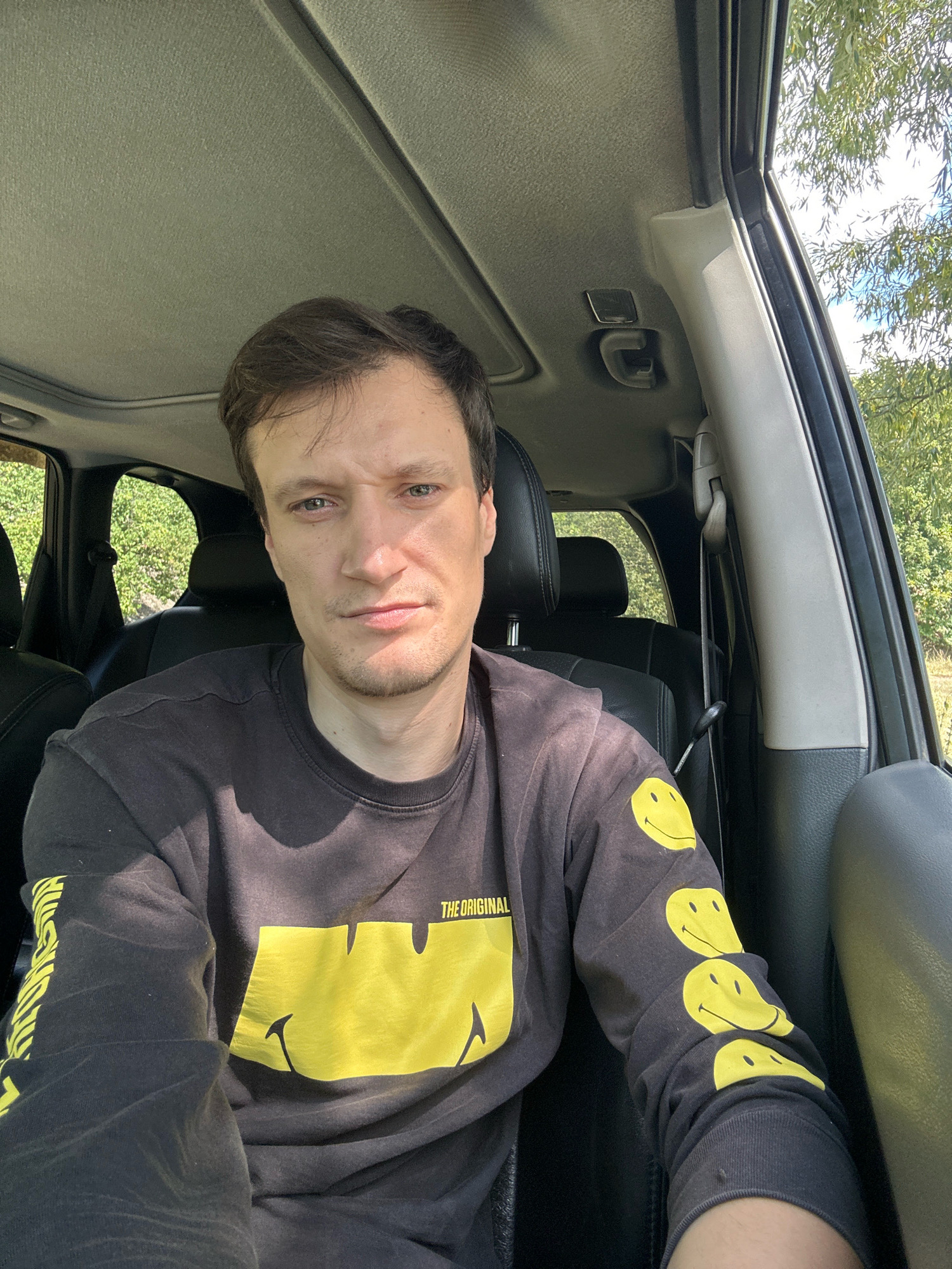 A male, appearing to be in his late 20s or early 30s, is seated in the driver's seat of a vehicle. He has light brown hair, light blue eyes, and is wearing a dark long-sleeved shirt with a large yellow smiley face design on the front and several smaller smiley faces on the sleeves. The vehicle's interior is visible, including black leather seats, the sunroof, and the car's dashboard. The background shows lush green trees and a bright blue sky, suggesting an outdoor location. The man is looking directly at the camera with a neutral expression. The location is identified as Makiv, Ukraine.