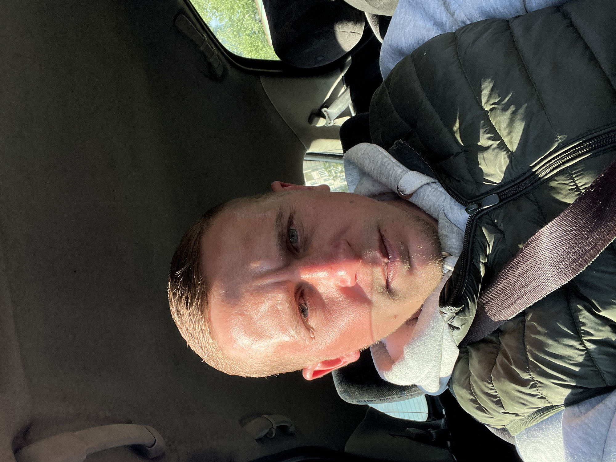 The image is a close-up, selfie-style shot of a person inside a vehicle. The person, who appears to be a young man with short blonde hair and blue eyes, is looking directly at the camera. He is wearing a grey hoodie underneath a dark green puffer vest. A seatbelt, with a textured purple strap, is visible across his chest. The lighting suggests it is daytime, with strong sunlight casting shadows on his face and vest. The background shows the interior of a car, including the car's headliner and a portion of the passenger seat. Sunlight streams in from a window, highlighting the person's face and the texture of his clothing. The overall mood of the image is casual and personal, likely taken by the subject himself. There is no visible text in the image. Given the provided location context of Minsk, Belarus, it is reasonable to assume the image was taken there.