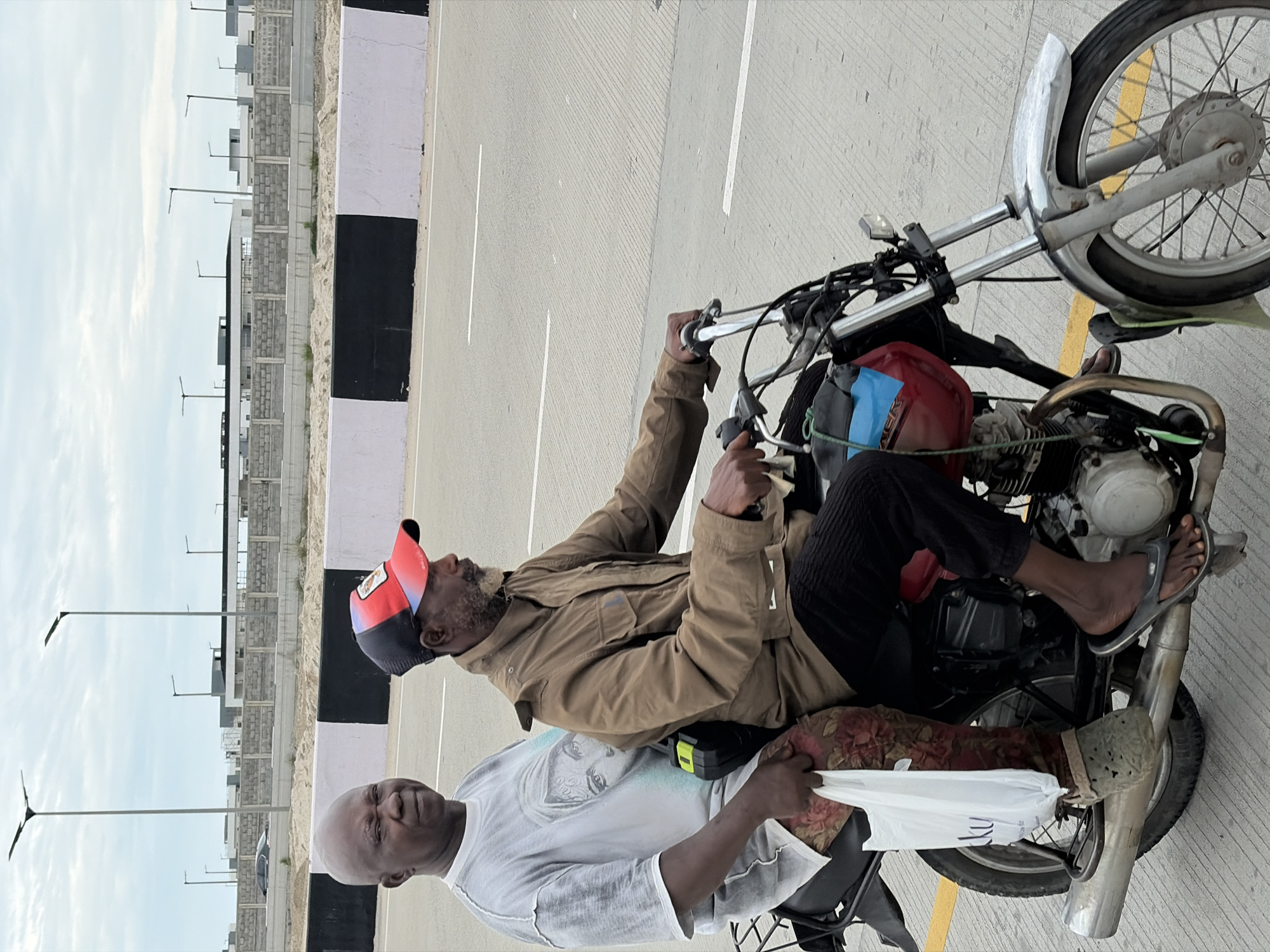 Two men are riding on a motorcycle on a paved road with white lane markings. The man in the front is driving and wearing a brown jacket and a red and black baseball cap. The man in the back is wearing a white t-shirt with an image of a face printed on it, patterned leggings, and grey clogs. He is holding a white plastic bag in his left hand. The motorcycle has a red and blue gas tank and a silver exhaust pipe. In the background, there are streetlights, a grey wall with black and white stripes, and buildings under construction. The scene appears to be in Igboefon, Nigeria.