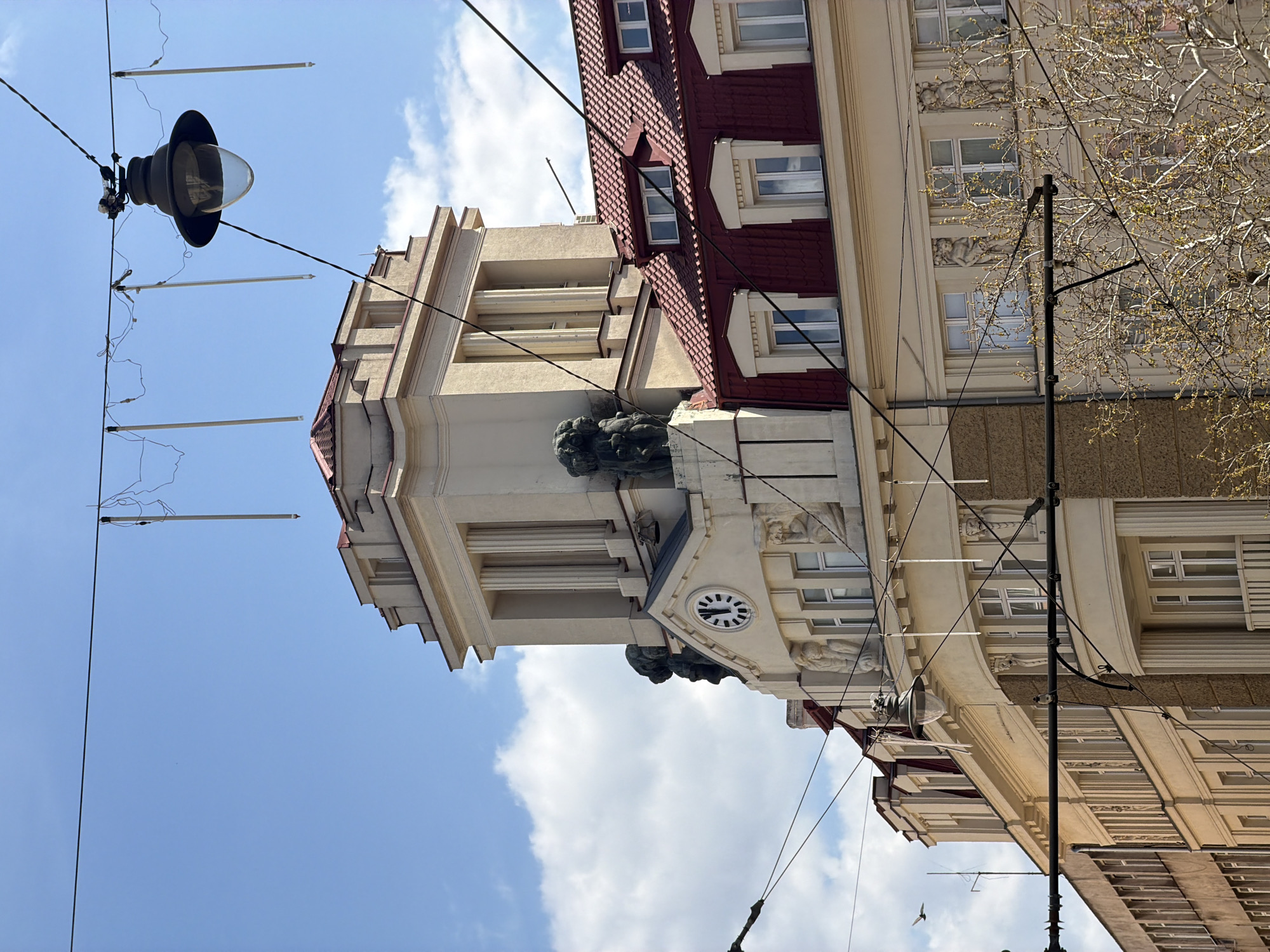 This is a brightly lit, upward-angled photograph capturing the architectural details of urban buildings and overhead infrastructure against a clear blue sky. The image, rotated 90 degrees counter-clockwise from its natural orientation, depicts a street scene in Sofia, Bulgaria, characterized by historical architecture and a network of overhead wires.
Two prominent buildings dominate the frame. The one on the left (when correctly oriented) is a light-colored structure featuring a distinctive tower-like section. This section is adorned with fluted pilasters or columns, intricate cornices, and dark, sculpted elements resembling gargoyles or mythical figures. A circular clock face, positioned below these sculptures, shows black hands indicating approximately 10:10. Adjacent to this, a prominent reddish-brown tiled mansard roof with multiple dormer windows adds to the building's character.
The building on the right showcases a facade decorated with intricate sculptural reliefs or friezes between its windows. Bare tree branches with nascent green leaves partially frame this building on the far right, suggesting the early spring season.
Numerous black wires, likely for electricity or public transport (trams/trolleybuses), crisscross the frame, with several white, tubular light fixtures suspended from them. A classic-style black streetlight with a clear glass dome hangs from these wires in the upper left corner. The sky is a vibrant blue with scattered white cumulus clouds, indicating a bright and sunny daytime. A small bird is visible in flight in the lower left portion of the image. No discernible text is visible apart from the stylistic marks on the clock face.