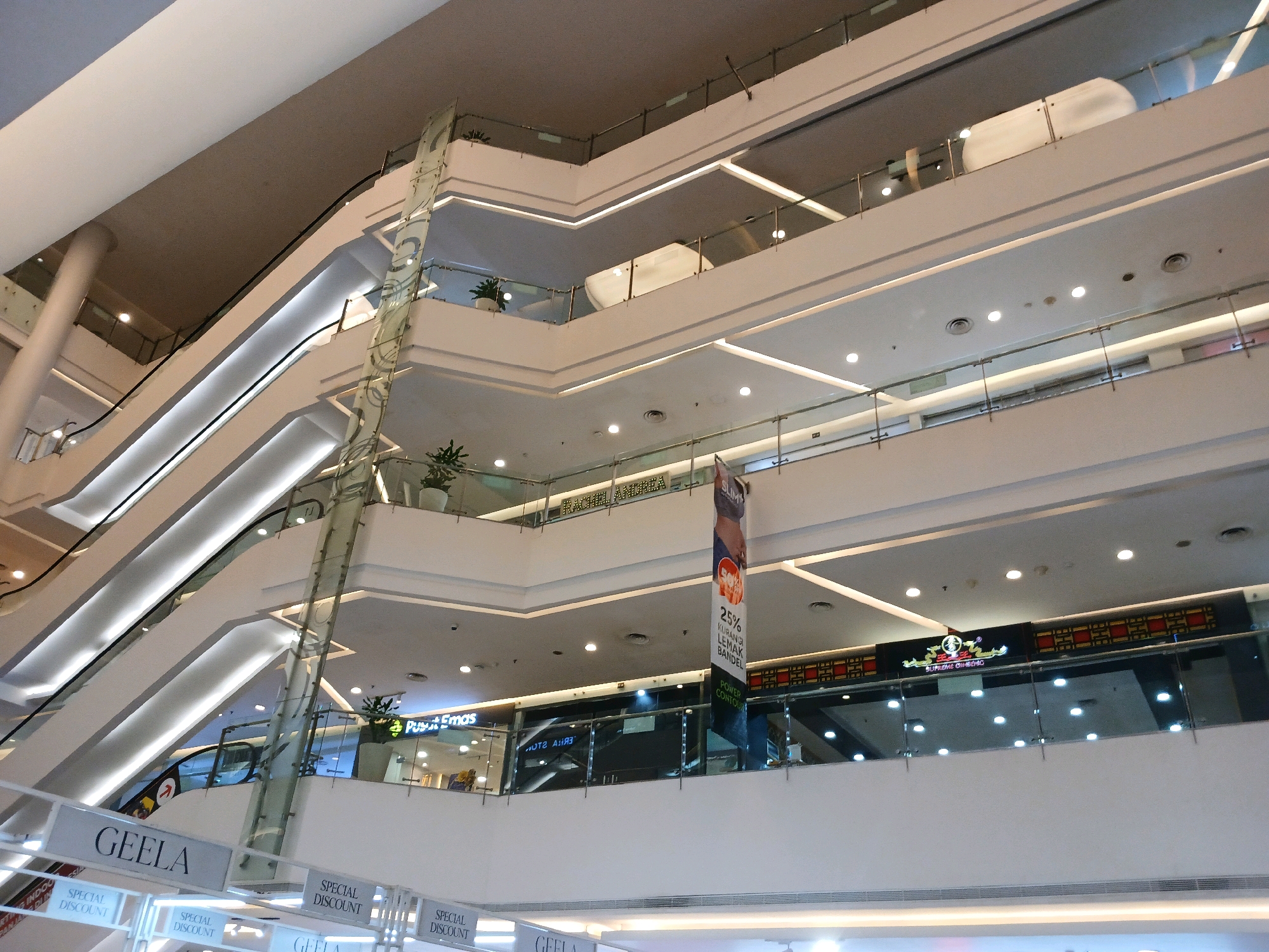 The image shows the interior of a multi-level shopping mall in North Jakarta, Indonesia. The architecture features a central atrium with escalators and walkways connecting different floors. The walls and ceilings are predominantly white and illuminated by recessed lighting, creating a modern and airy atmosphere. Several shops are visible on different levels, some with prominent signage like "GEELA" and "Pusat Emas." A banner advertising a "25% KURANGI LEMAK BANDEL POWER CONTOUR" is displayed in the atrium. The scene is observed from a lower level looking upwards, capturing the layered design of the mall's interior. There are no discernible people in the frame.