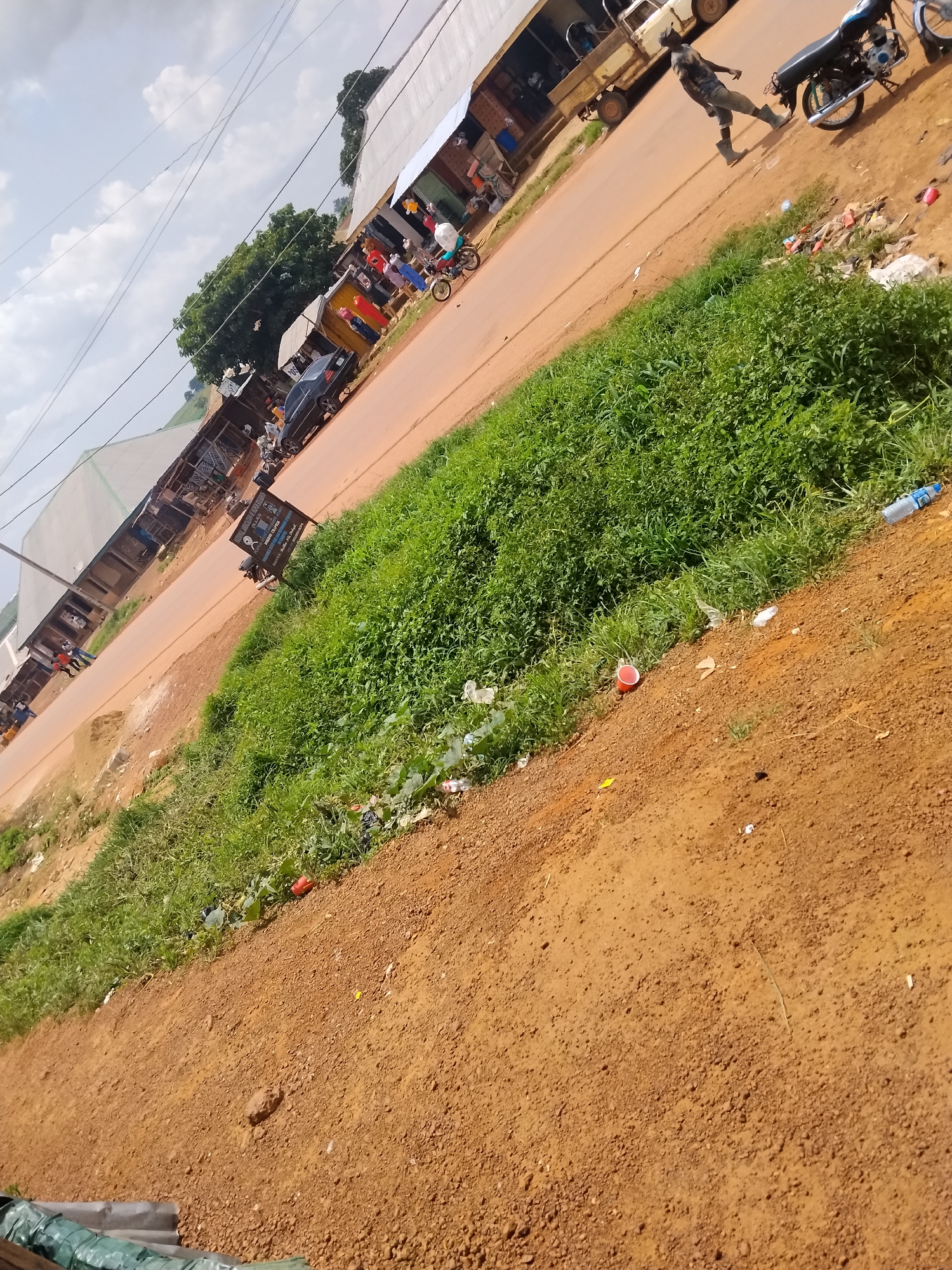 The image depicts a street scene in Durumi, Nigeria. The scene is characterized by a dirt road, a grassy embankment, and several small shops. A person is walking on the road, and a motorcycle is parked nearby. There are also several discarded items, including plastic bottles and cups, scattered on the ground. The sky is cloudy, and the overall atmosphere suggests a casual, everyday setting.